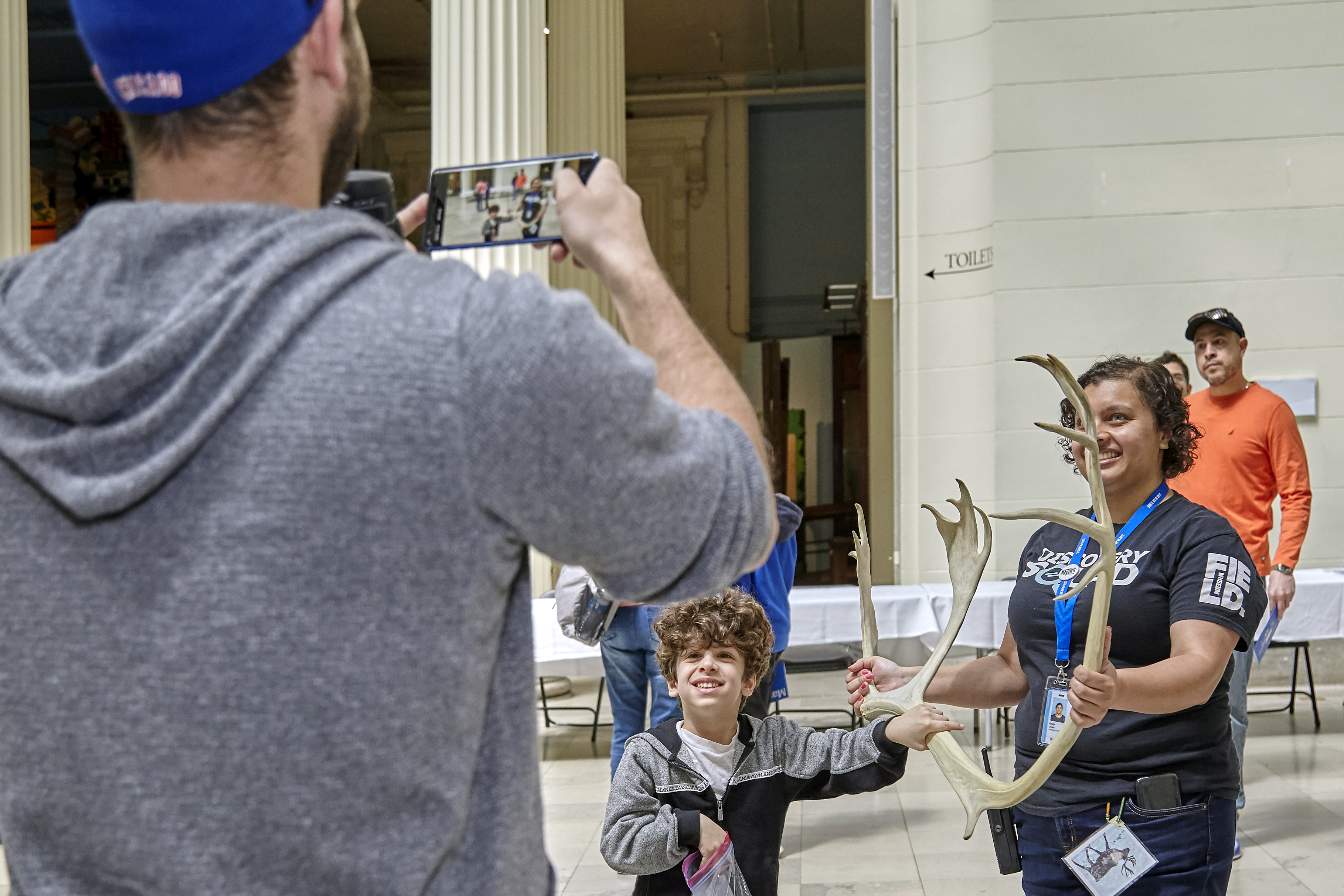 a museum volunteer stand with a young visitor, holding a antler, while an adult visitor takes a photo