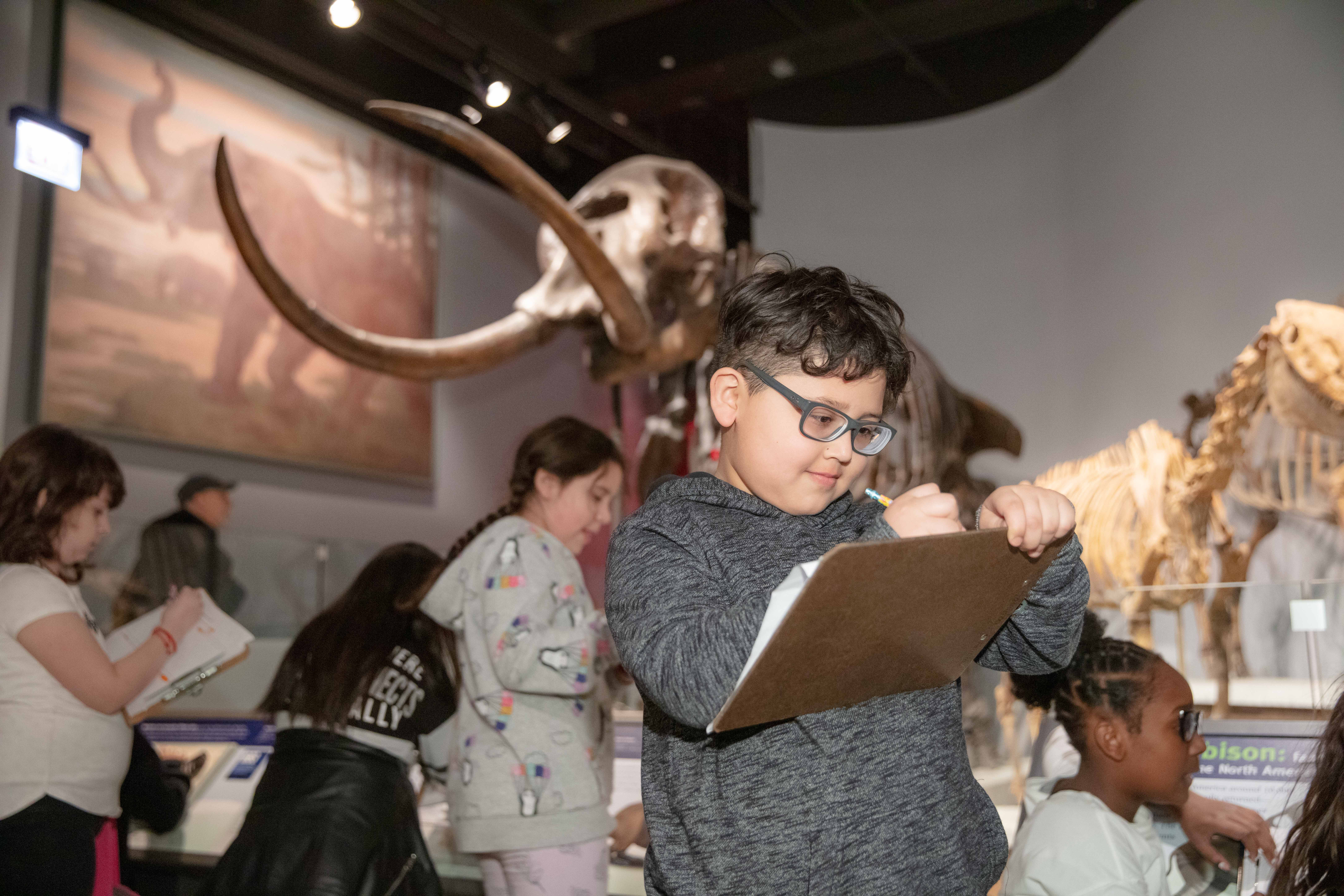 A young student writes on a clip board he is carrying through a museum exhibition. Other students and fossils can be seen in the background.