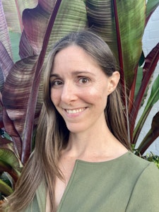 A woman with long brown hair, wearing a green top, stands in front of a large plant.