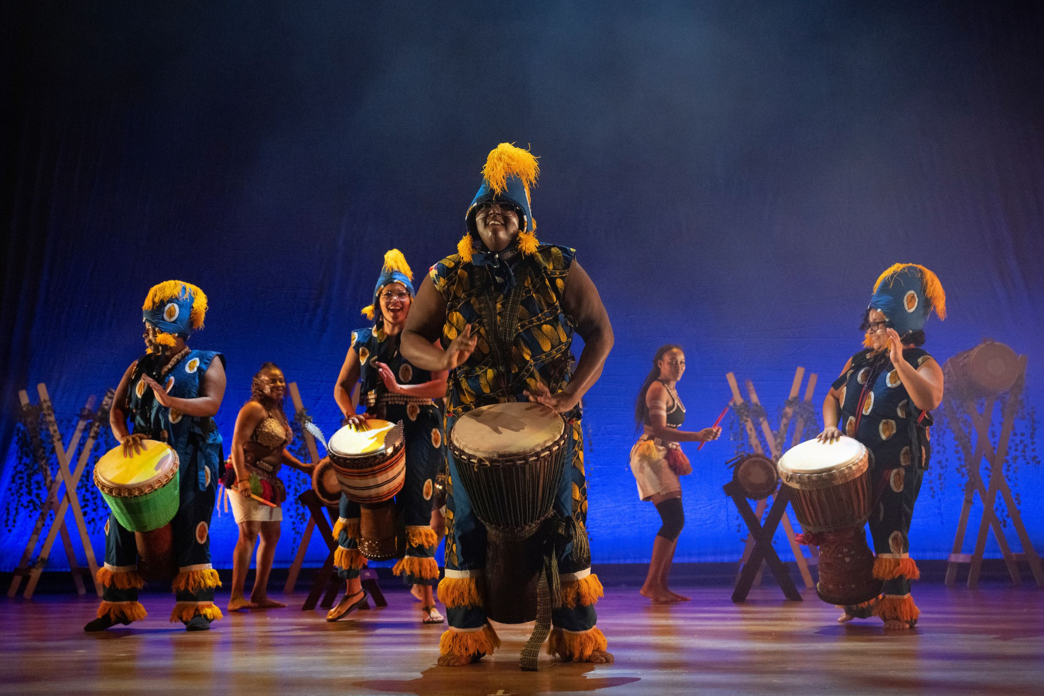 Several performers wearing traditional African regalia dance and play instruments including drums