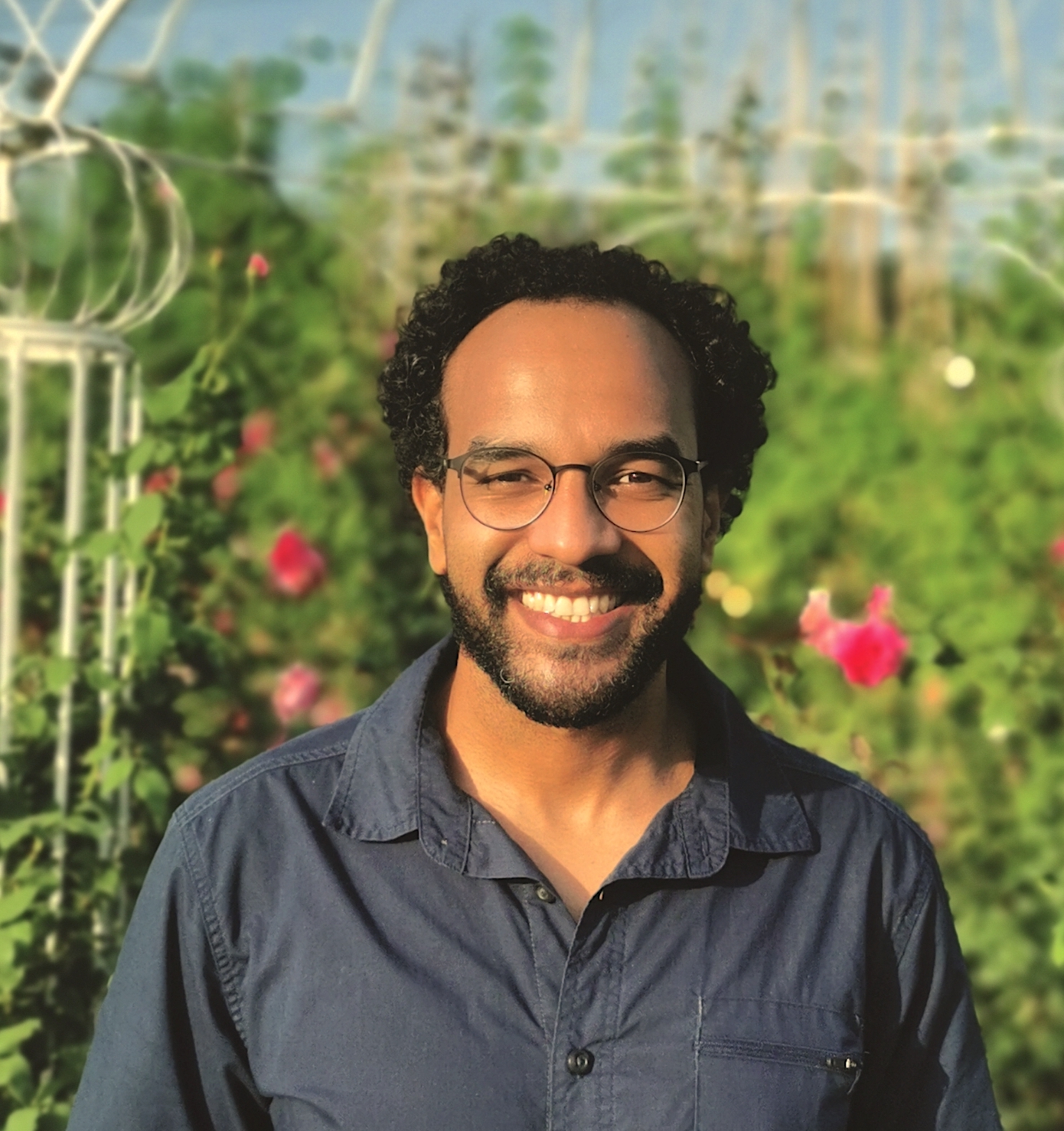 A man with black curly hair and glasses stands smiling in front of a garden.