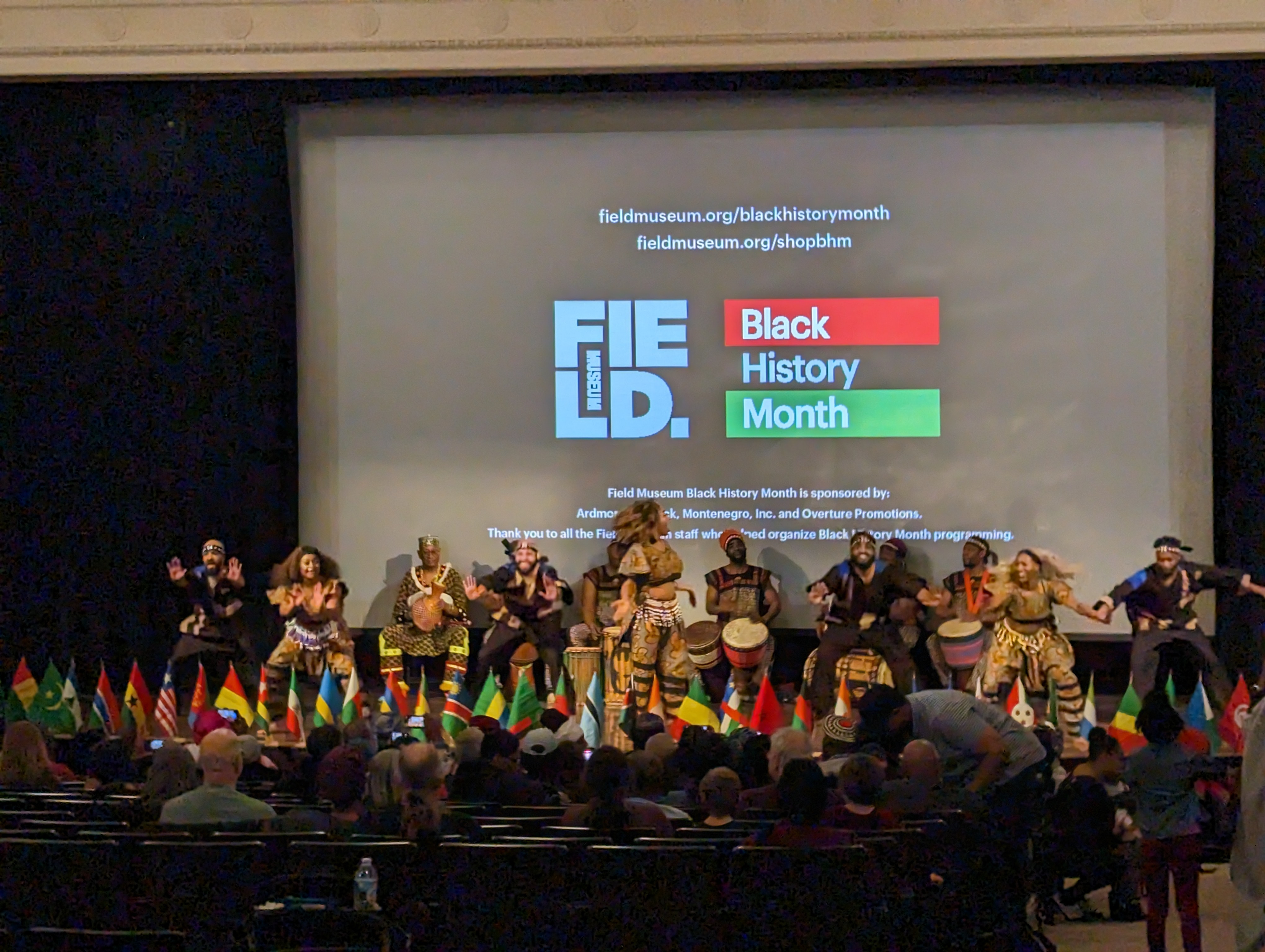 a group performs on a stage in front of an audience. A large screen behind the performers shows Field Museum and Black History Month logos.