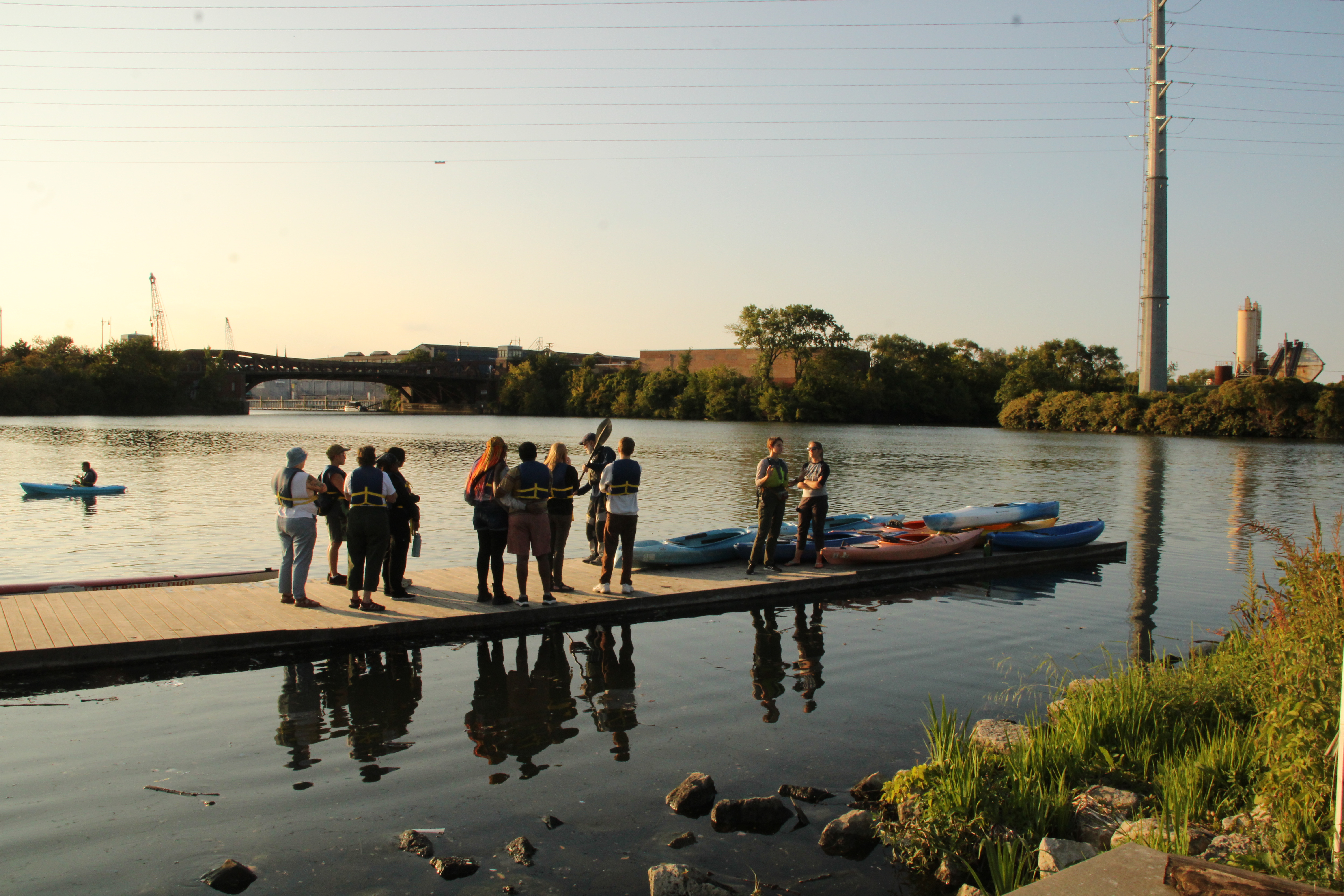 A group about a dozen people stand on a pier on a river, with kayaks staged nearby. It appears to be sunset.
