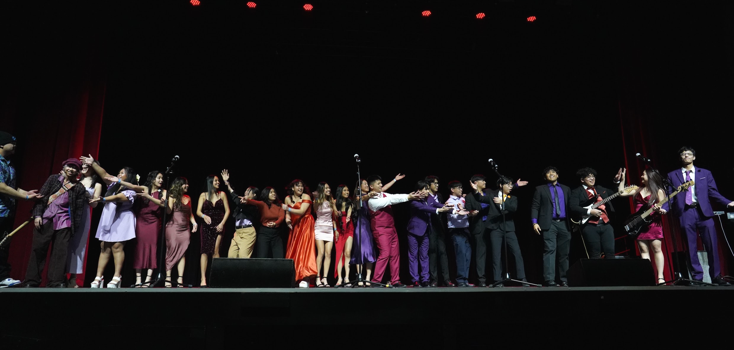 A group of young people, dressed semi-formally, stand together on a stage. Some of them are holding musical instruments.