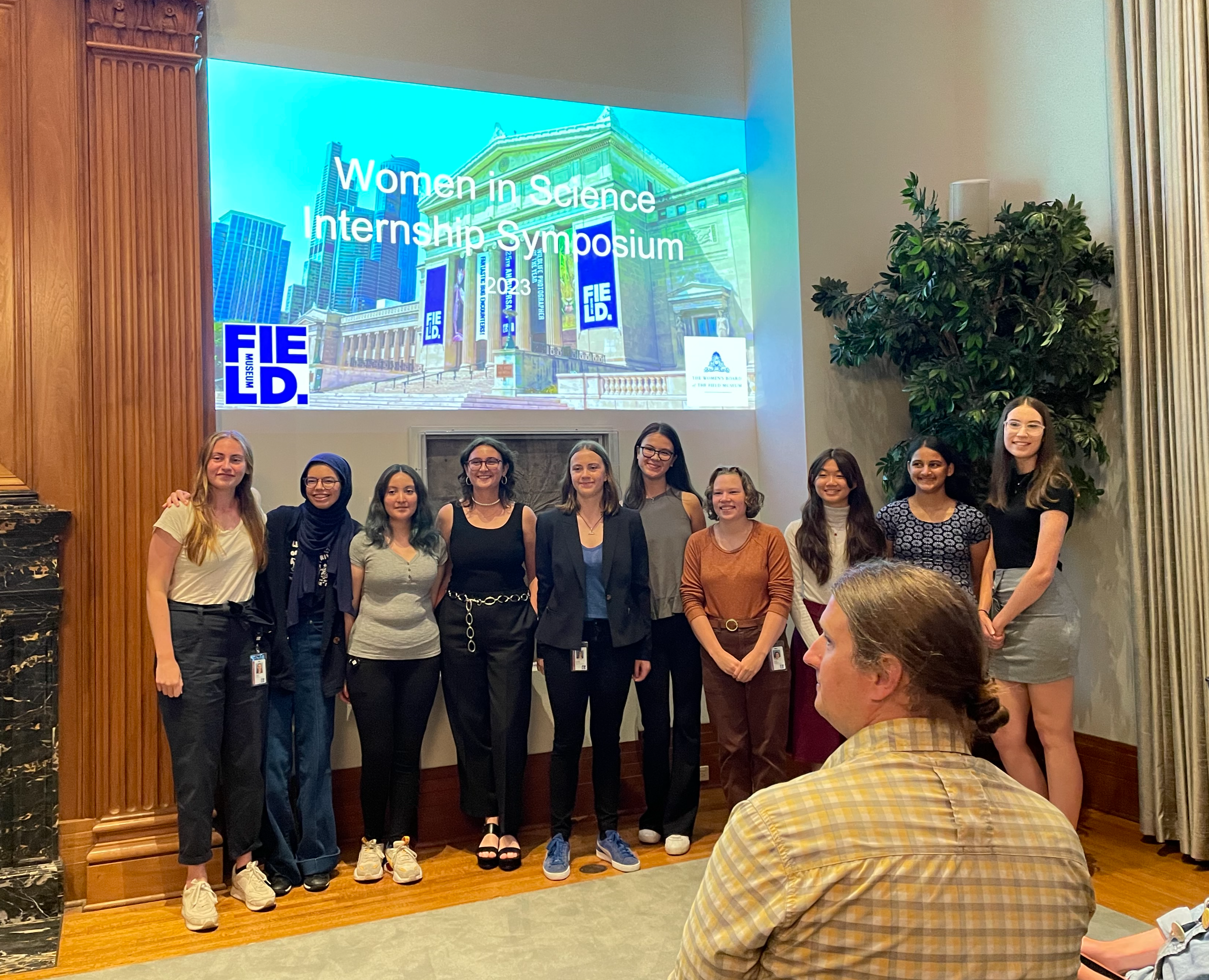 A group of young women stand below a video screen at the front of a presentation room.