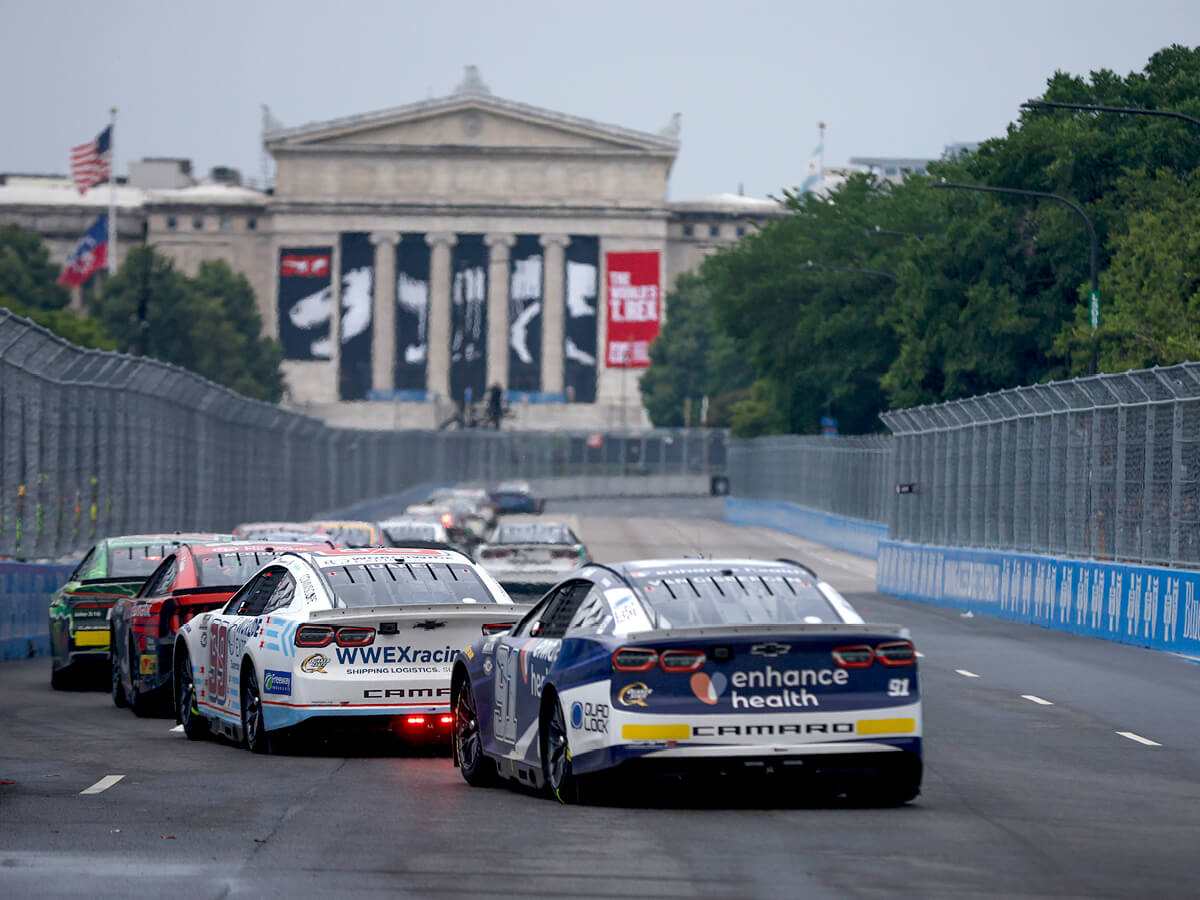 Several stock cars racing together on a road course. The north facade of the Field Museum can be seen in the background @NASCAR