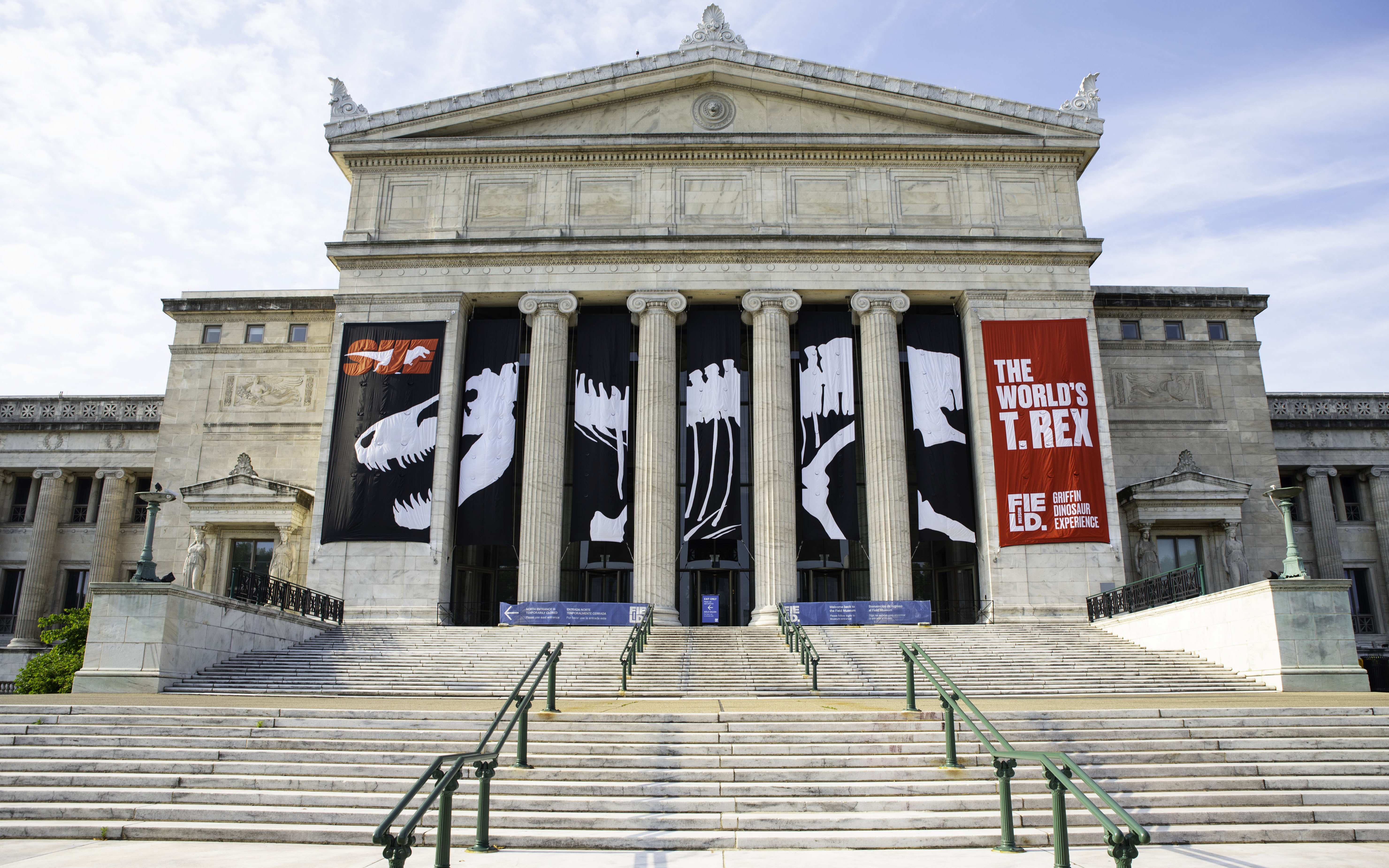 A large, classical building, with steps leading up to four ionic columns. Black, white and red banners are displayed on the building.