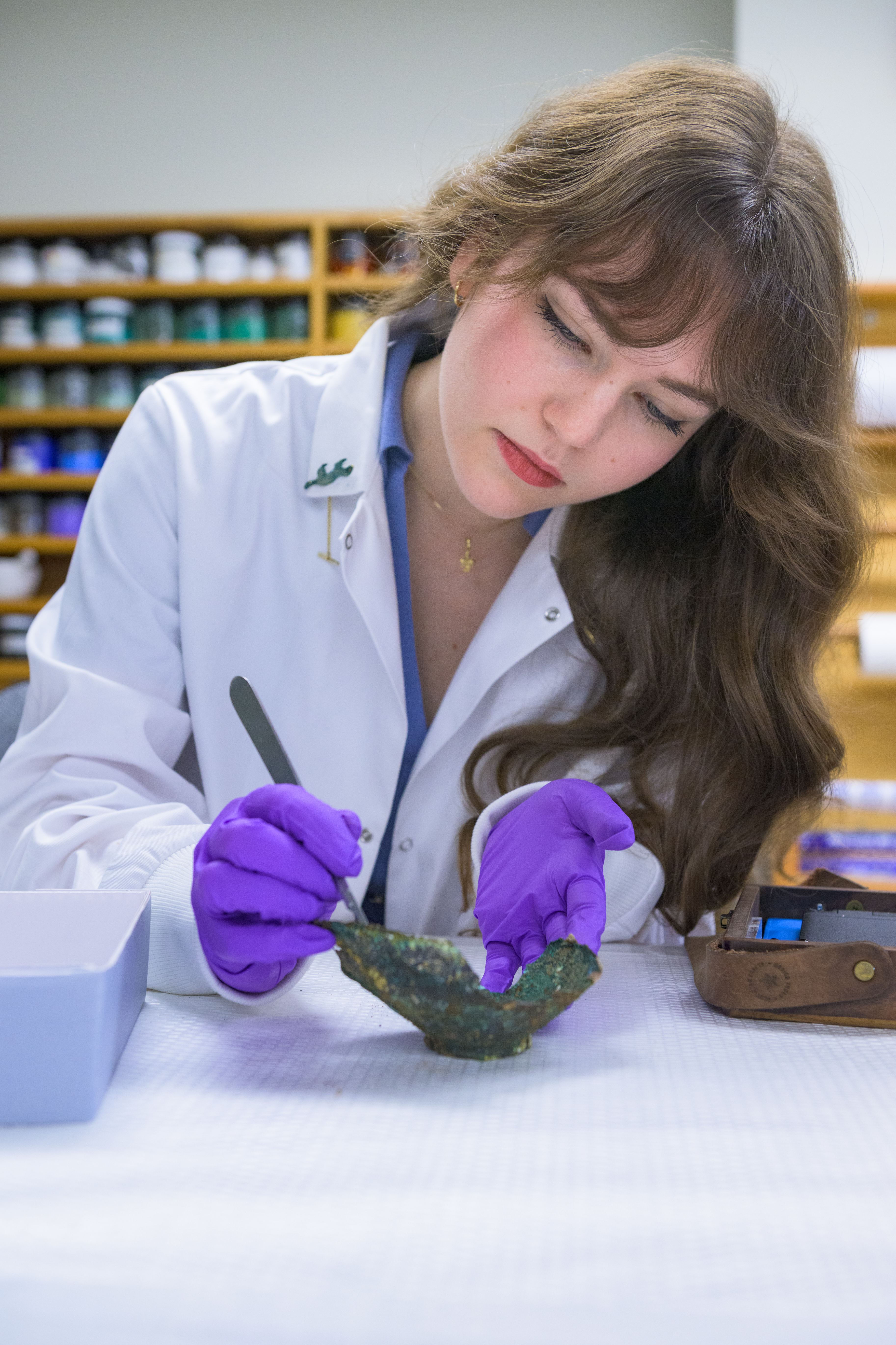 A woman wearing a lab coat and exam gloves. She looks down at a specimen as she examines it with a tool.