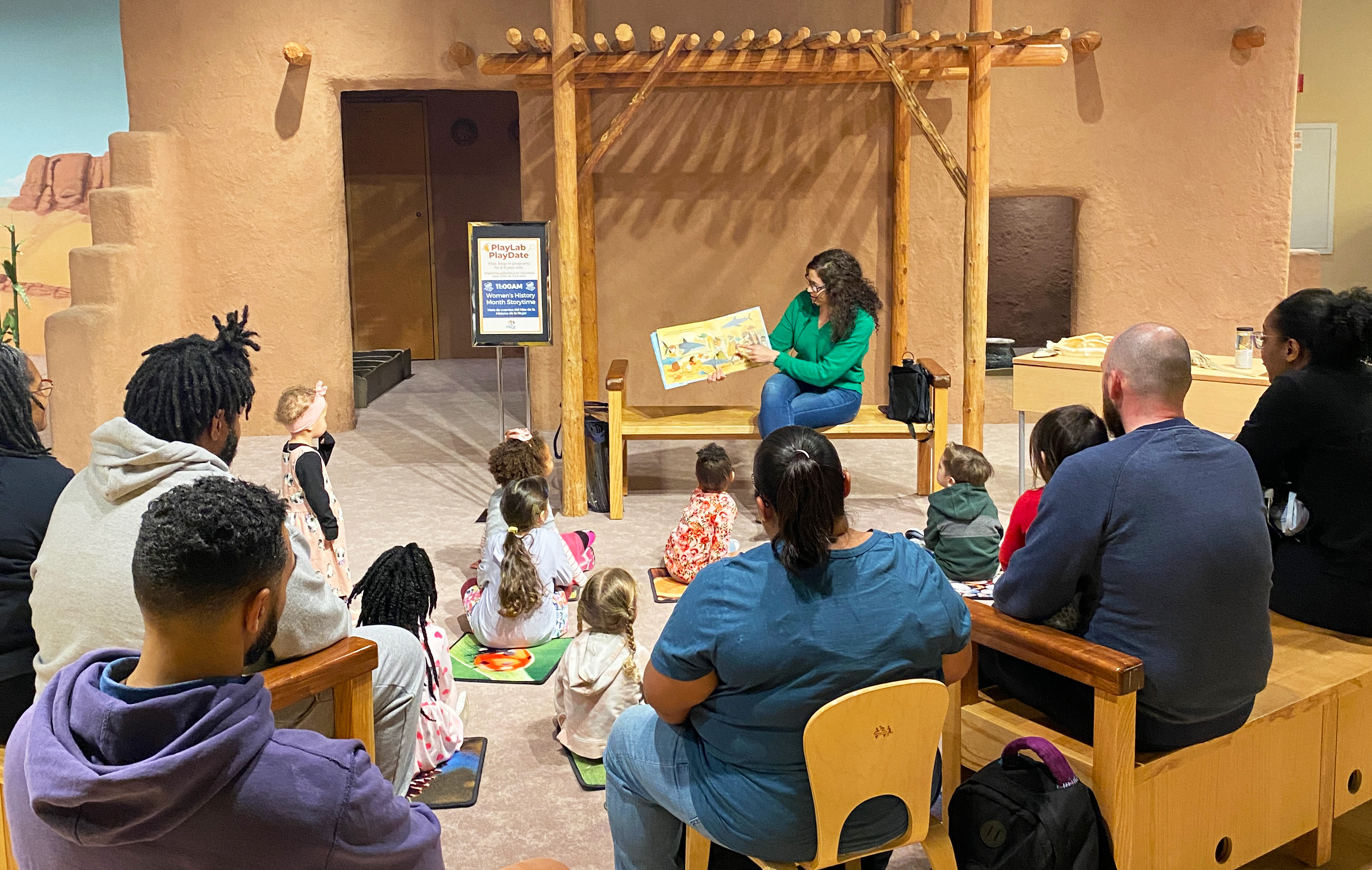 An educator sits at the front of a group holding a book for them to see. Children are seated on mats on the floor as adults sit on chairs, all facing the reader.