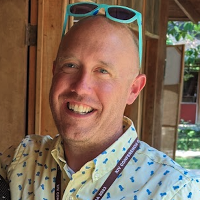 A man, smiling and wearing a button down shirt. His sunglasses are perched on top of his head.