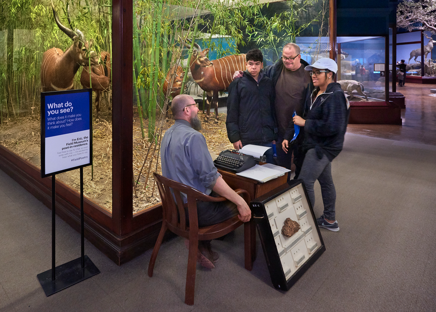A group gathers at the corner of a diorama case. One man sits a desk with a typerwriter. He looks up to a family of three standing over him. The man in the middle stands with arms around the other two individuals. Both are smiling down at the man seated at the desk. A black box with a glass front holds a rock specimen and is propped up against the leg of the desk.