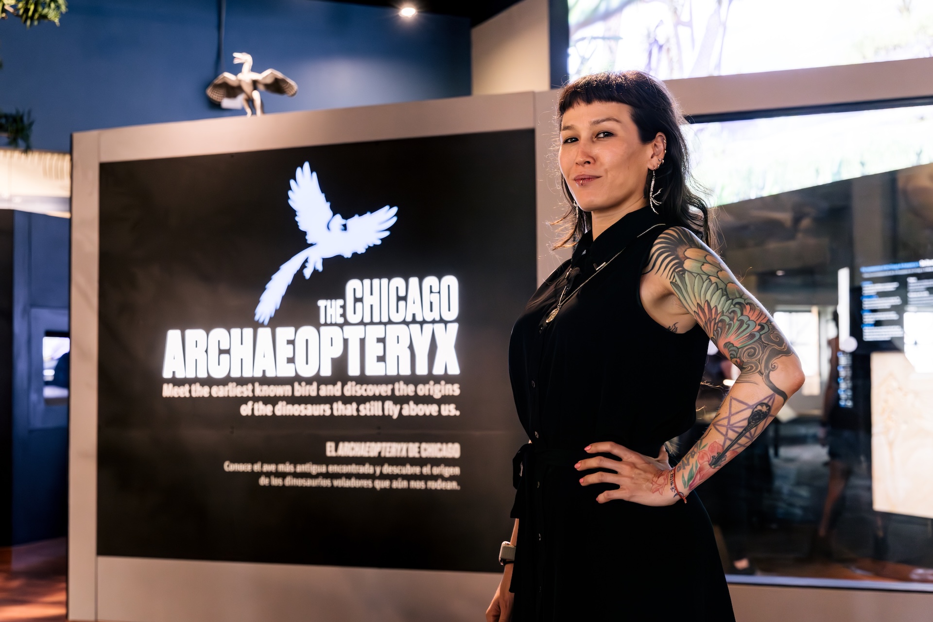 A woman stands next to a graphic panel in the Chicago Archaeopteryx exhibit.  She is wearing a sleeveless black dress with her tattooed left arm rests on her hip.