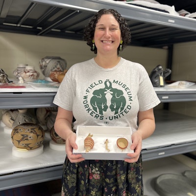 A woman wearing a t-shirt holds a box with three museum specimens as she stands in front of shelves.