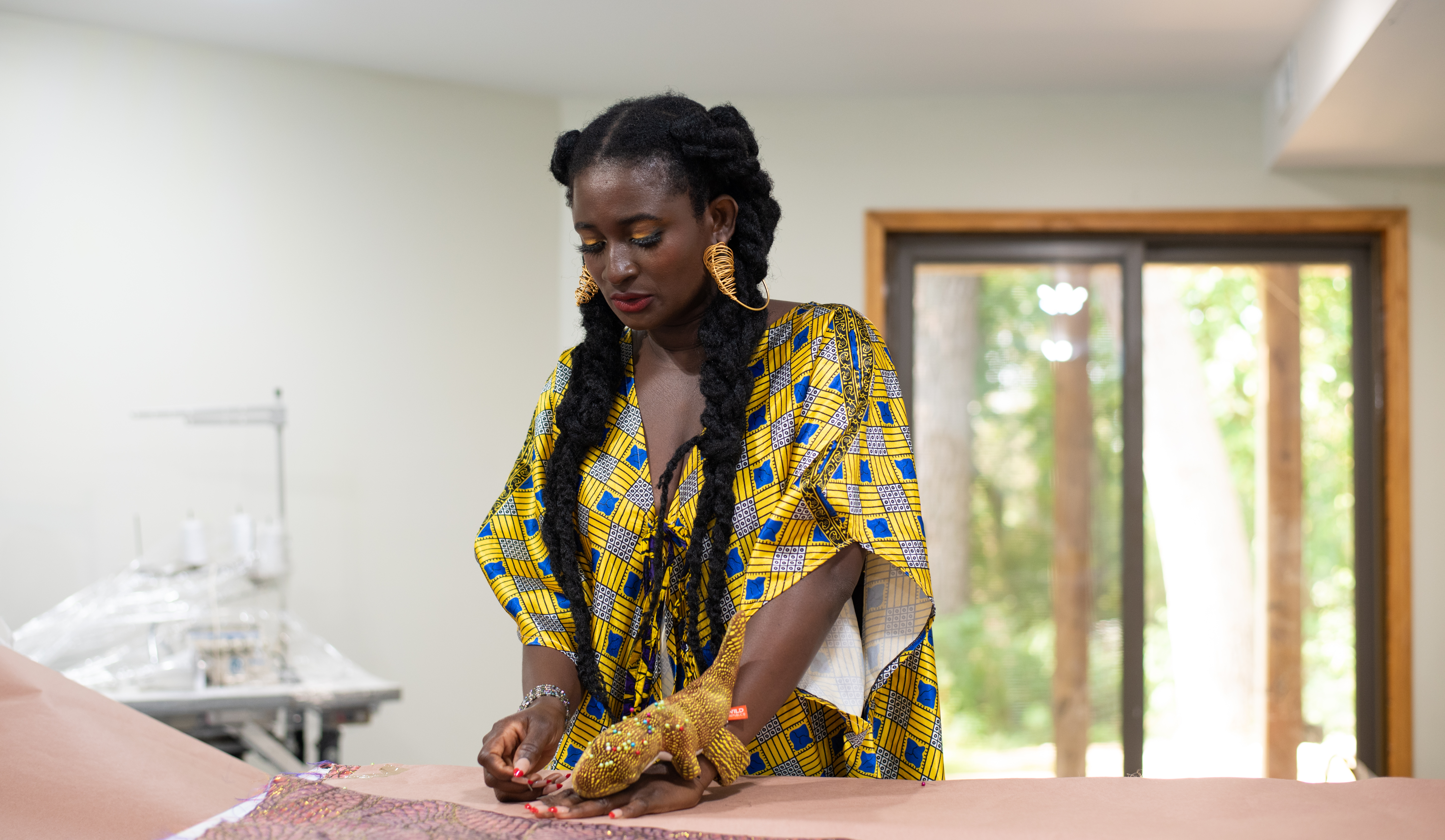 A woman with long black braids, gold hoop earrings, and a blue and yellow patterned dress works in her studio.