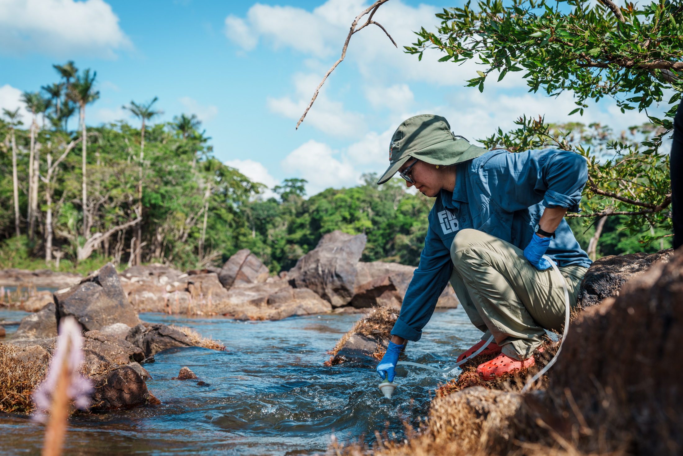A person wearing a sun hat and rubber gloves stoops down, reaching into a river with testing equipment.