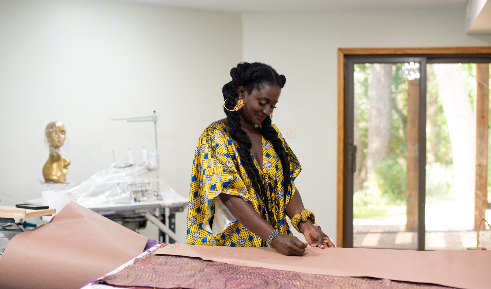 A woman with long black braids, gold hoop earrings, and a blue and yellow patterned dress works in her studio.