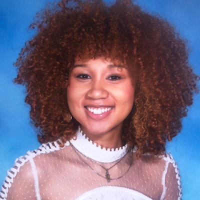 A woman with brown curly hair, wearing a white high-neck top.