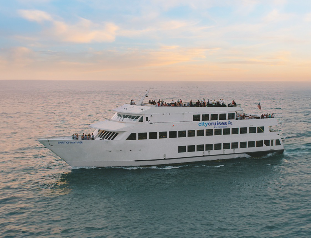 People can be seen on the outside decks of a large yacht cruising on calm waters, pink clouds in the sky.