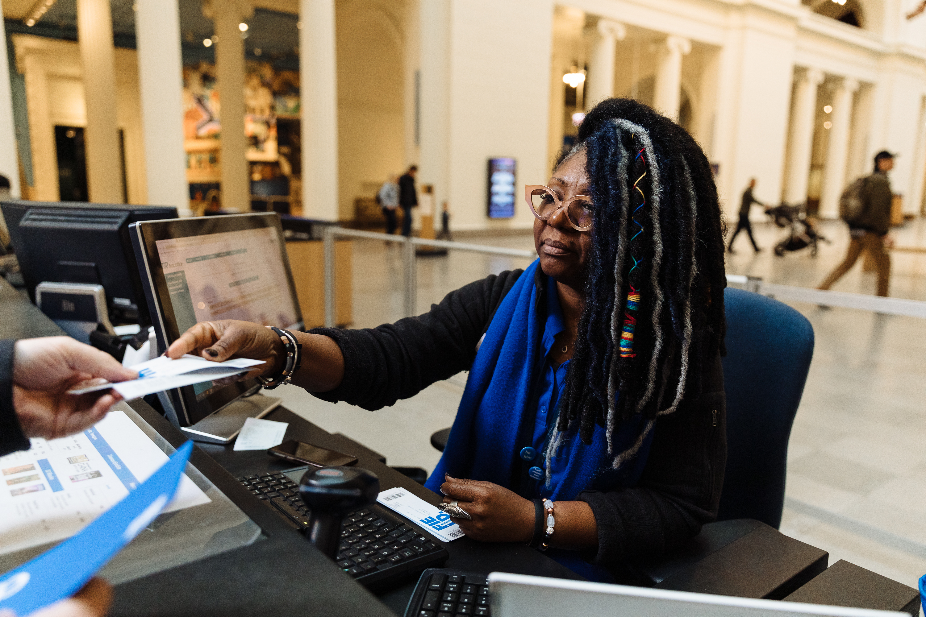A woman seated at a counter with a computer and keyboard hands papers to a person with only their hand visible.