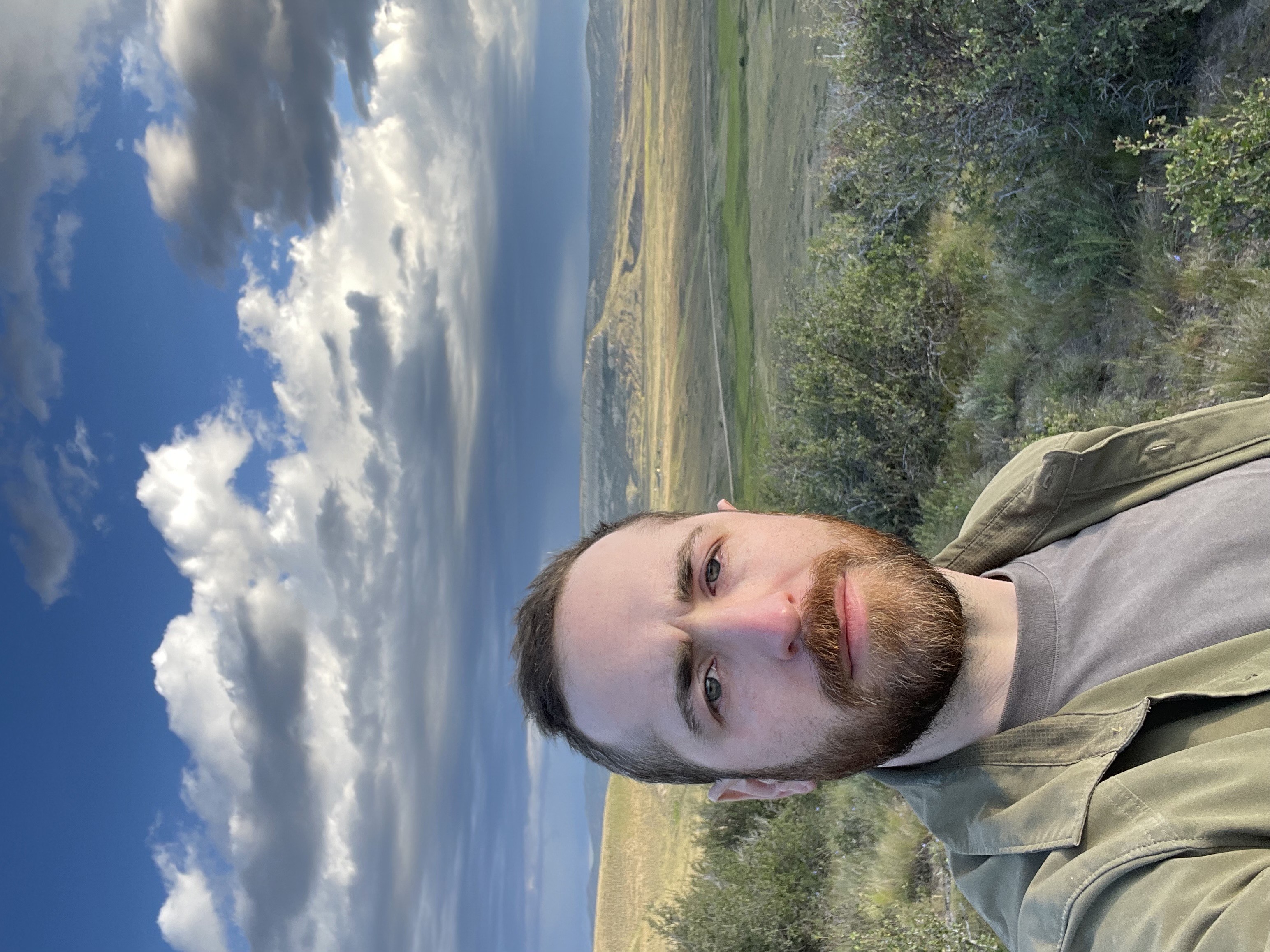 A man stands in front of an open landscape with rolling grassy hills. White and grey clouds fill the blue sky.