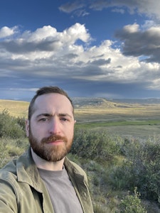 A man stands in front of an open landscape with rolling grassy hills. White and grey clouds fill the blue sky.