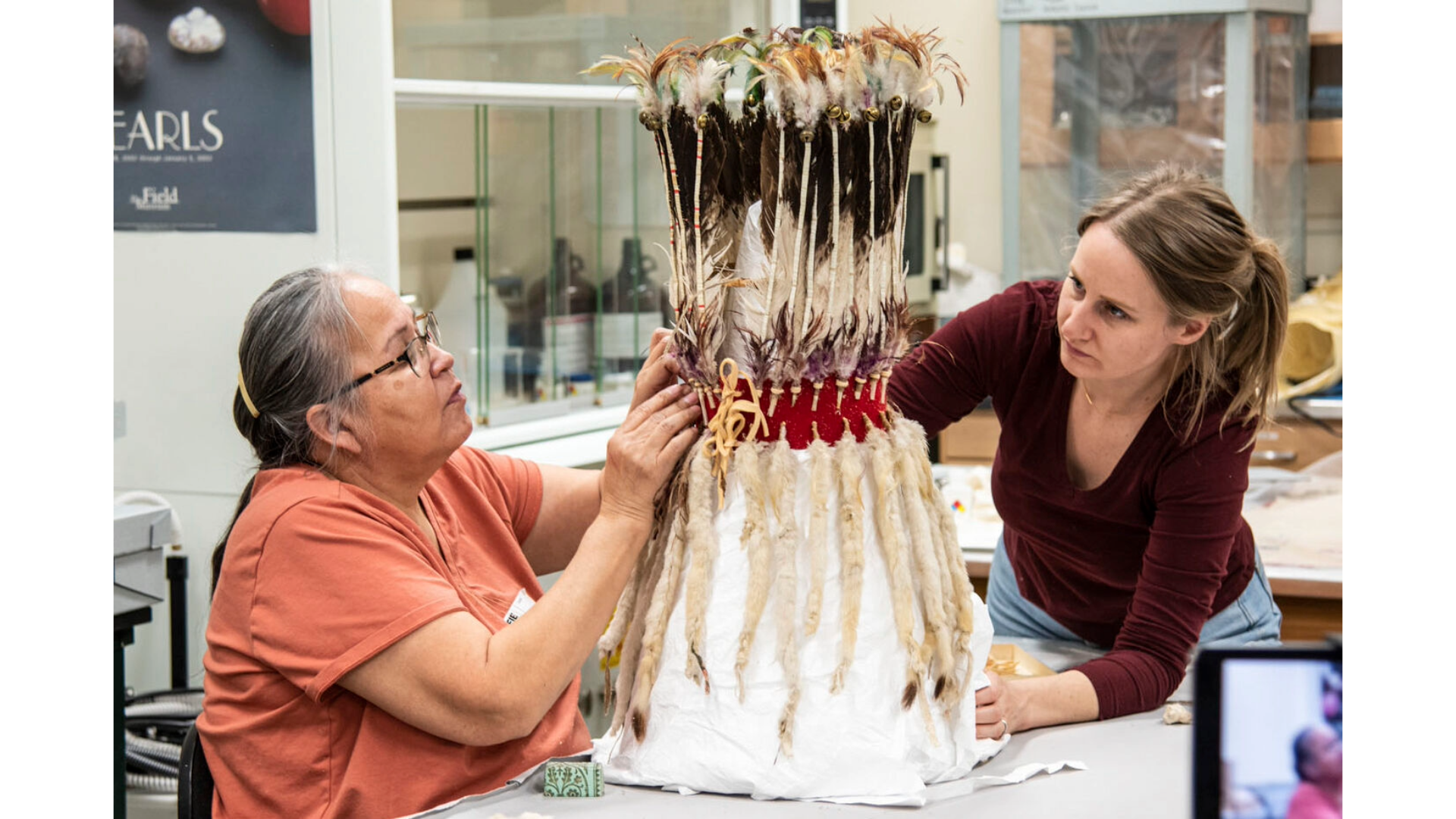 Two women leaning toward a stand-up headdress sitting on a table in a lab.