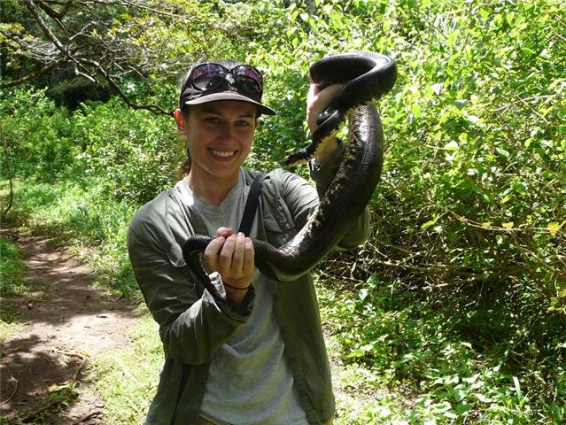 A woman holding a large brown snake in her hands stands in a wooded path.