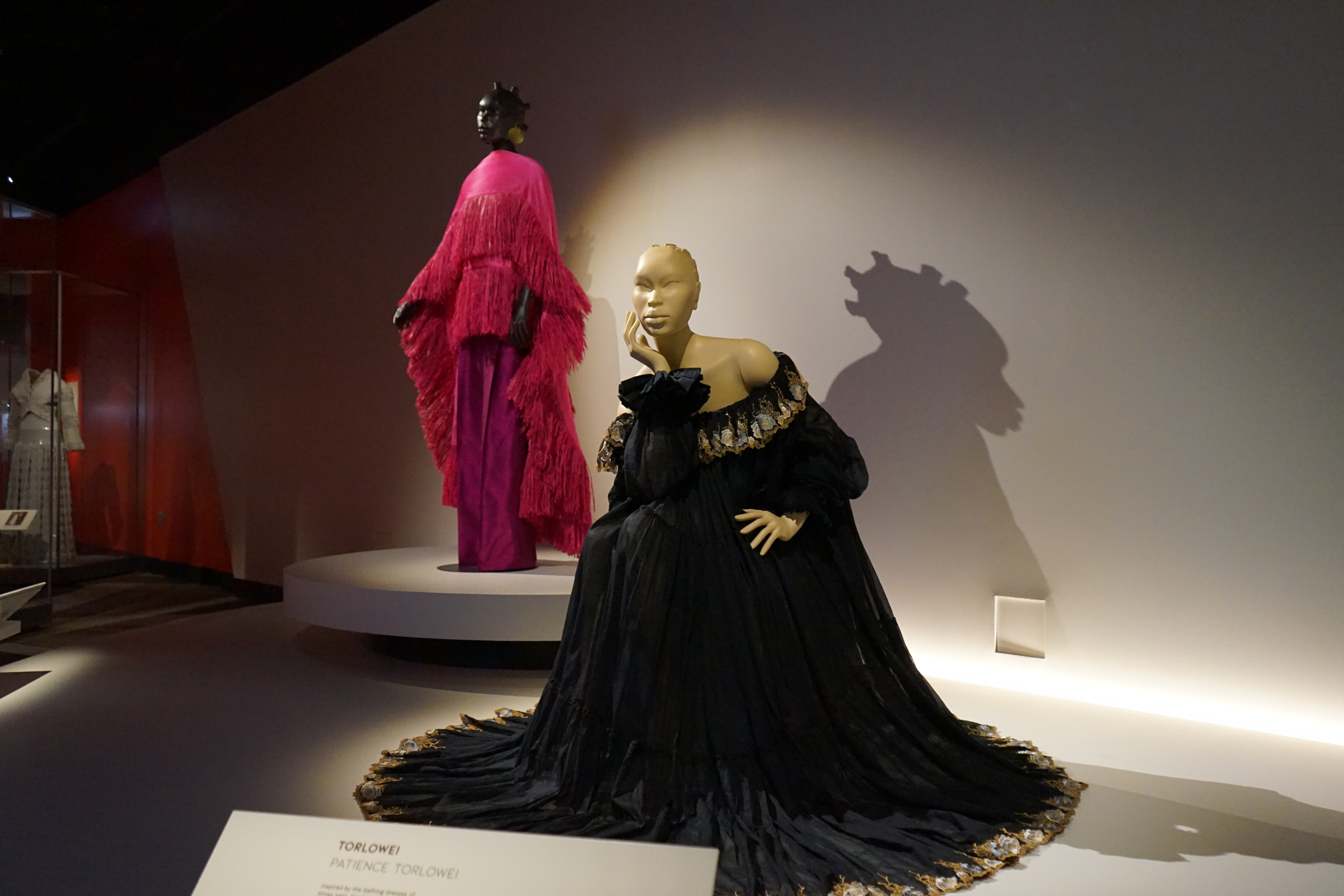 A black dress on a seated mannequin, with another mannequin standing in the background wearing a bright pink dress.