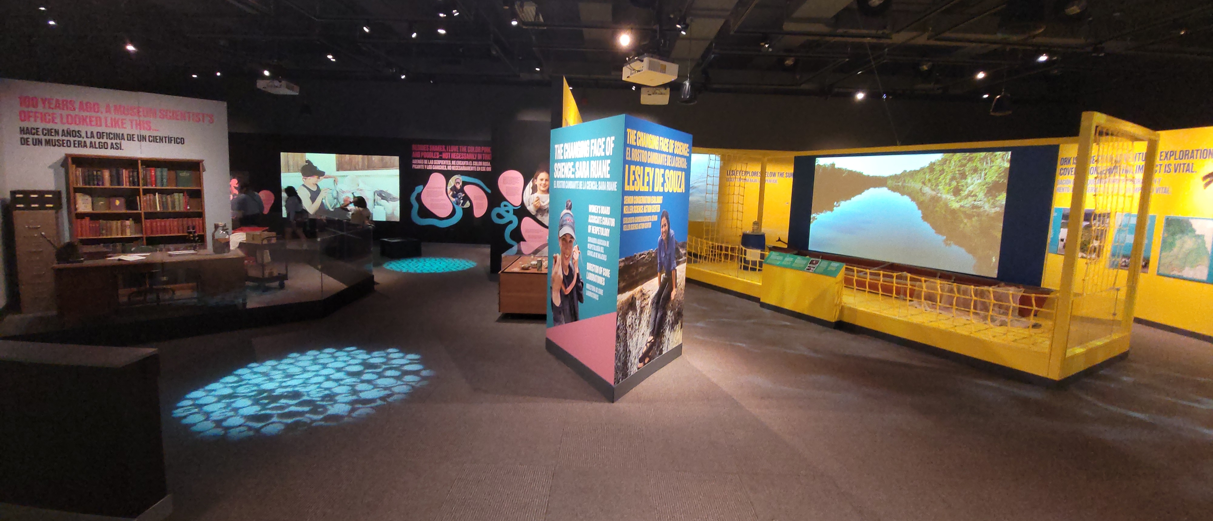 A panoramic view of two museum galleries with large video screens, graphic panels and objects displayed.
