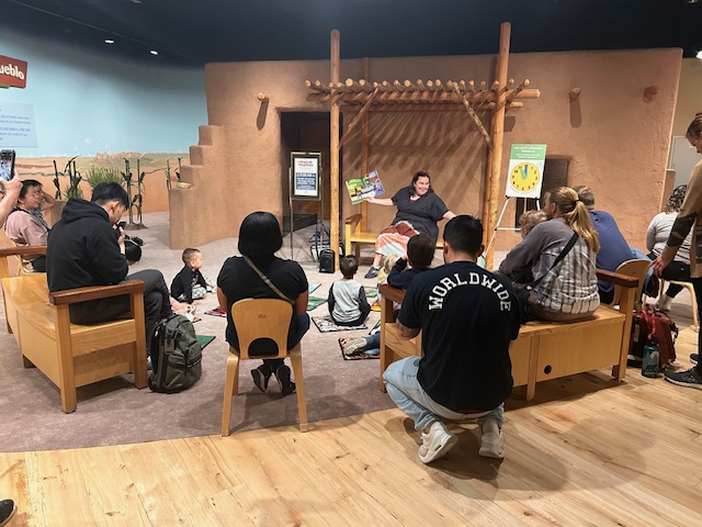 A woman sits holding a book, facing a group of parents and children.