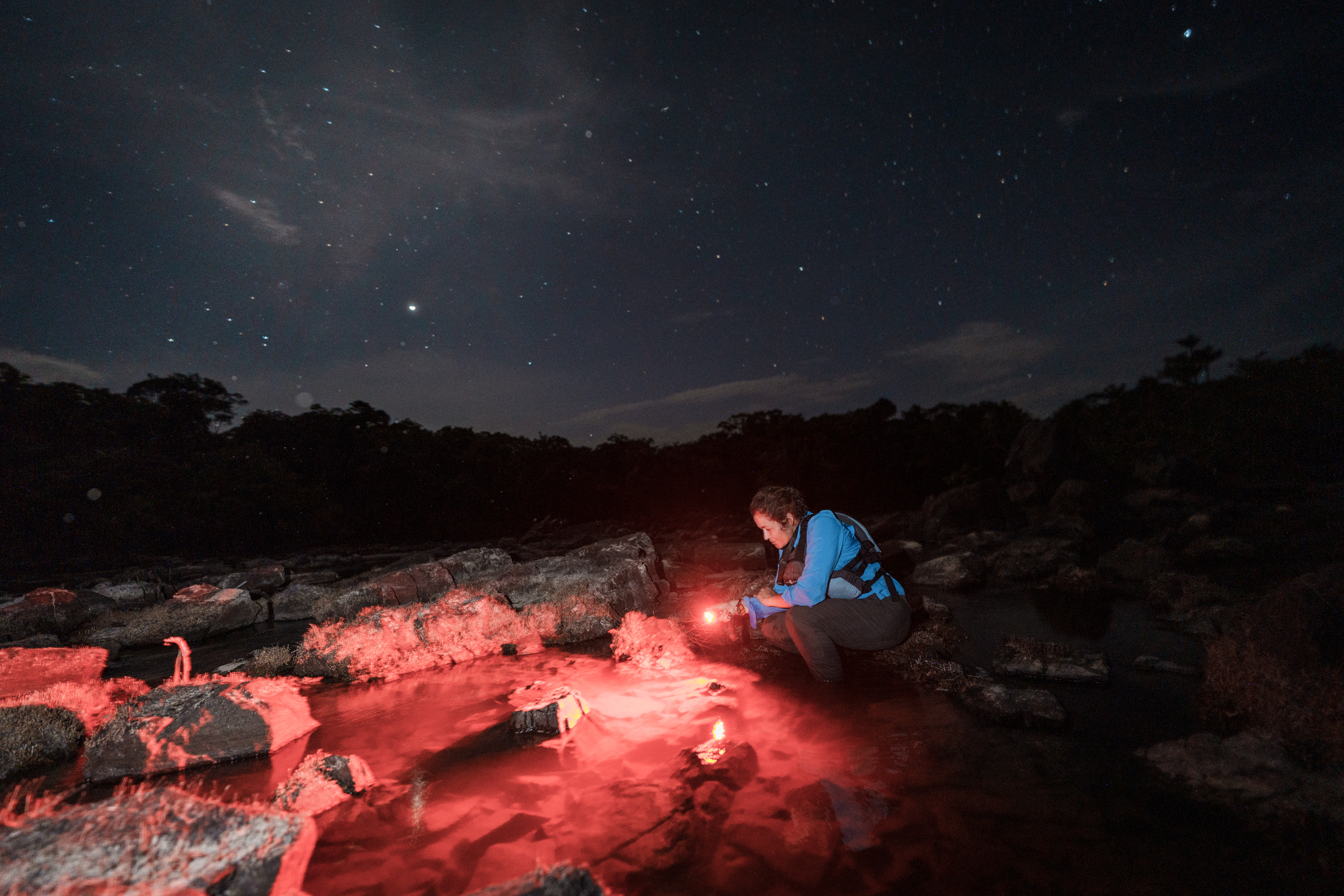 A person stooped on a rocky surface with red flares lighting the area. A dark night sky shines above.