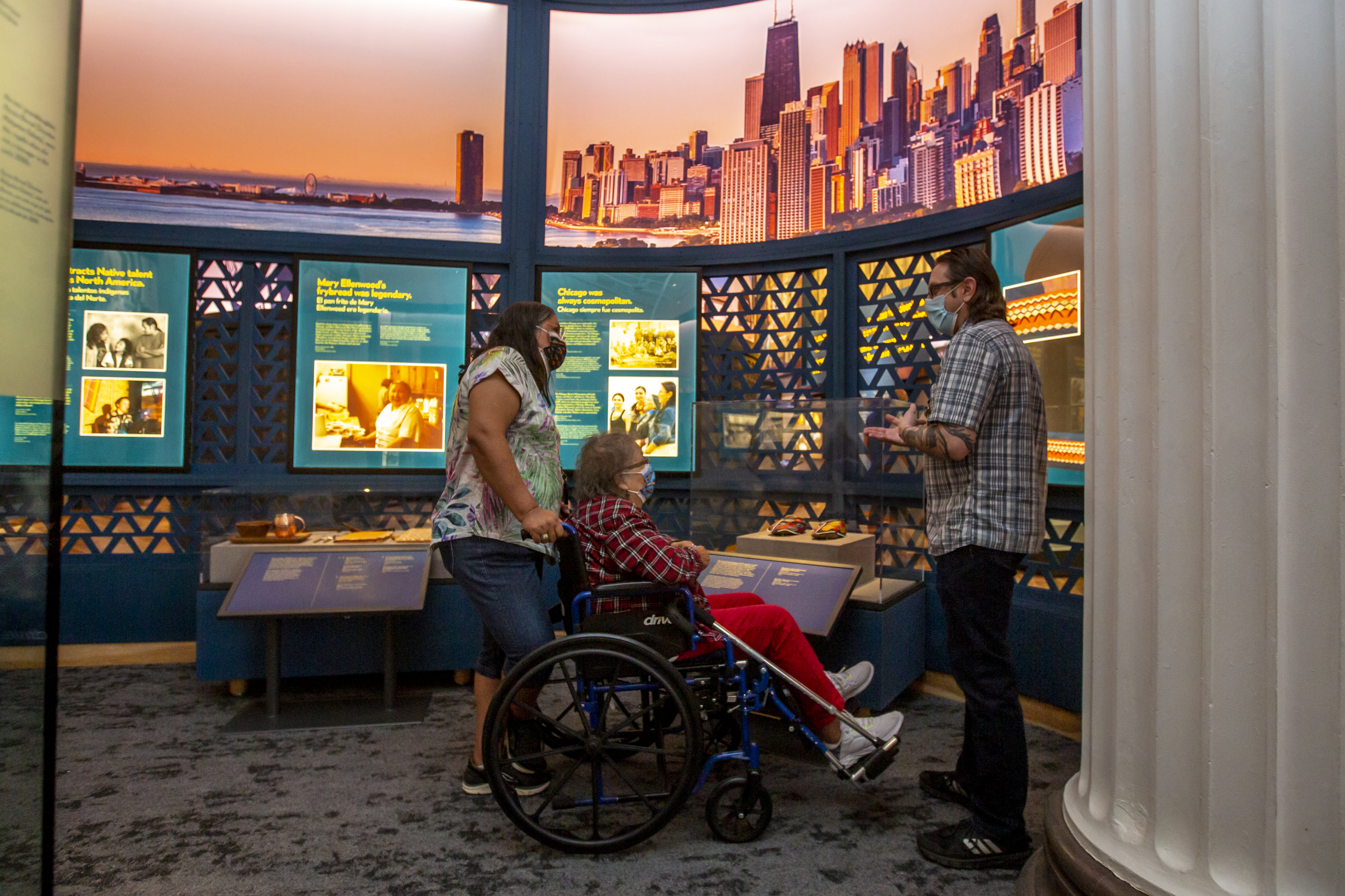 A person seated in a wheelchair, being pushed by another person. Both are facing a third person in a museum exhibition with graphic panels around them.