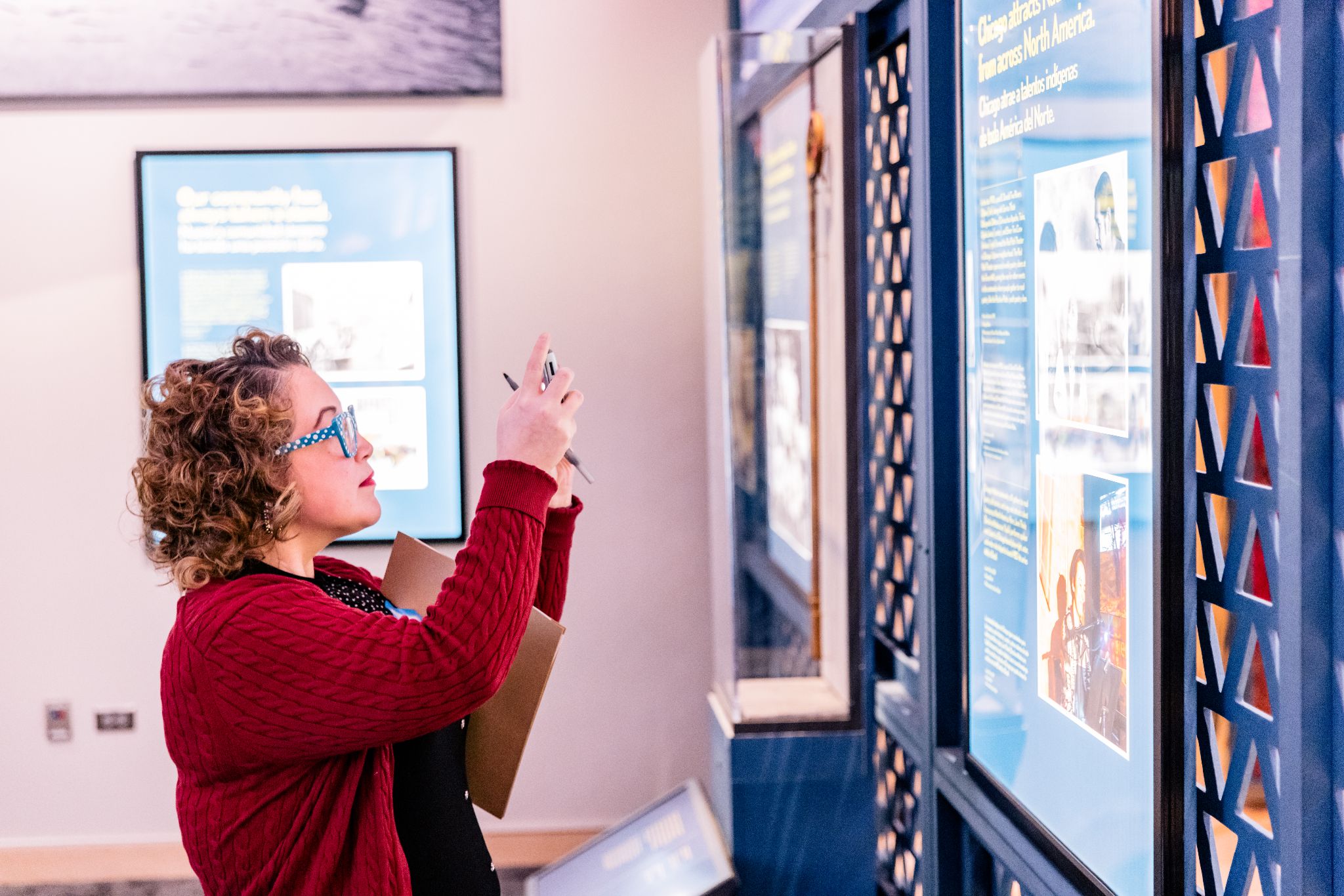 A woman wearing a red sweater and glasses holds her phone up to take photos of graphic panels in a museum exhibition.