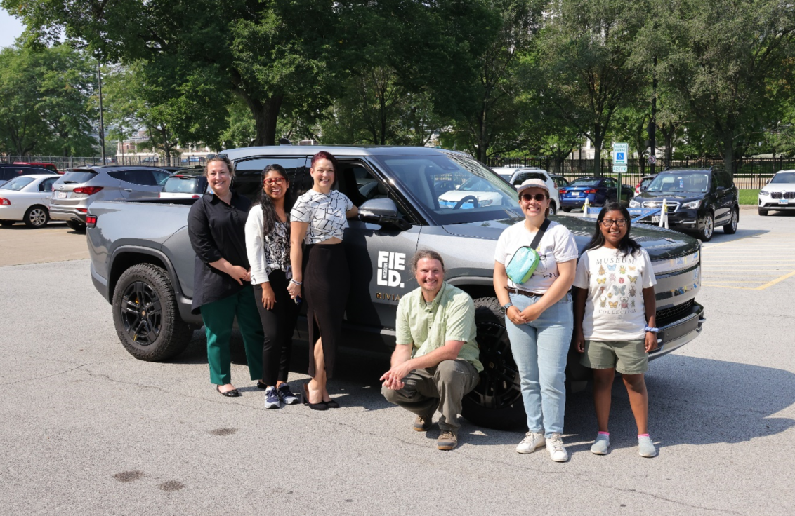 Six people stand next to a grey pick-up truck in a parking lot.