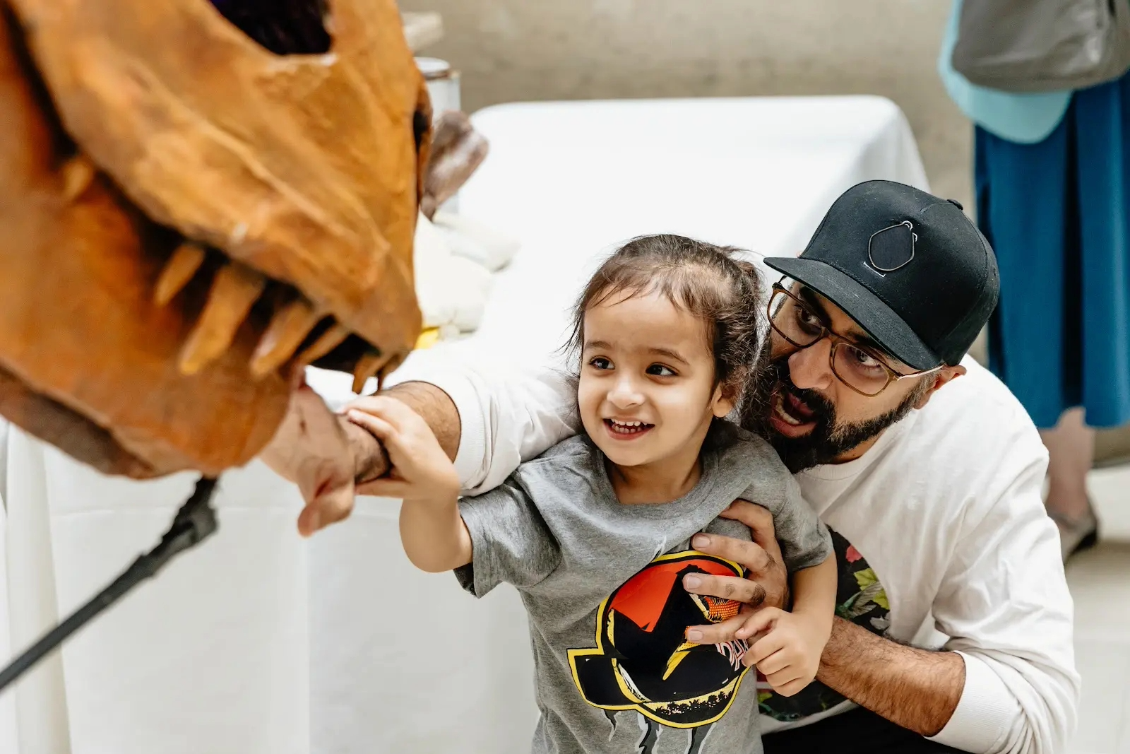 A man stoops down behind a child as they reach out toward a dinosaur puppet, partially seen.