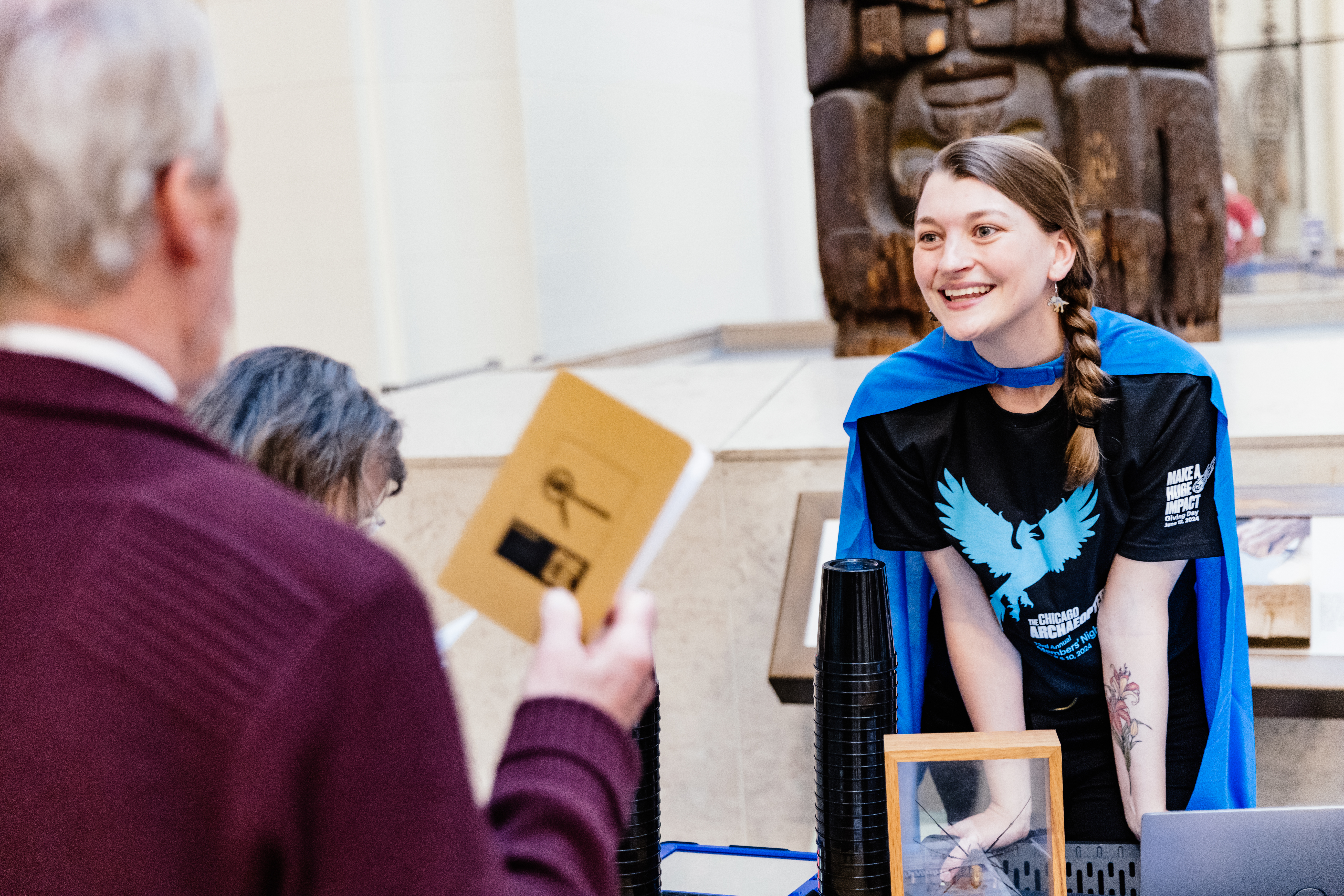 A volunteer interacts with visitors in Stanley Field Hall. The volunteer is smiling broadly and wearing a bright blue cape and a Chicago Archaeopteryx t-shirt.