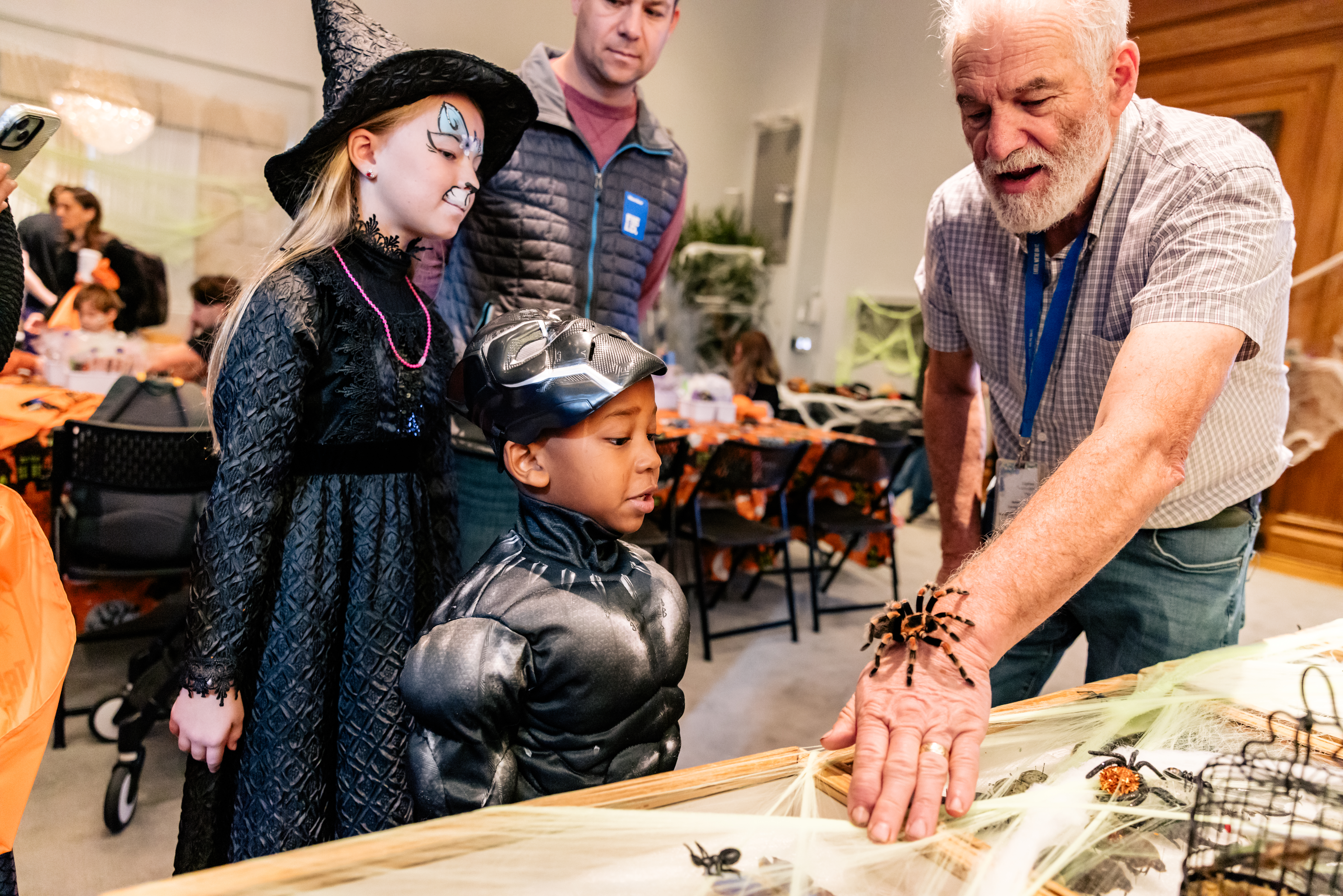 Museum staff member extends his arm as a tarantula crawls on his wrist. Museum visitors in halloween costumes look on.