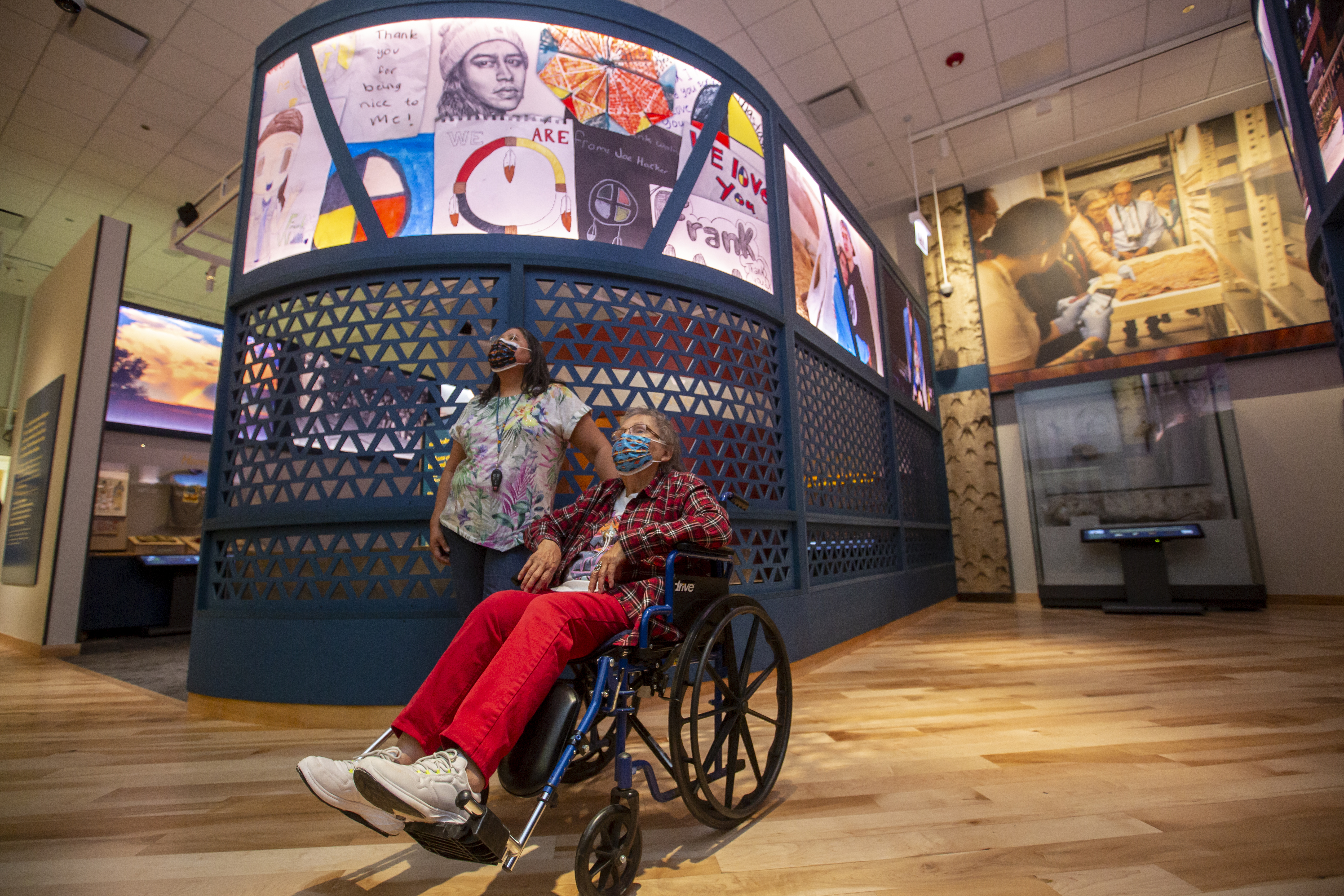 Visitors inside the Native Truths: Our Voices, Our Stories exhibition look up at an exhibit. Both visitors are wearing medical face masks. One sits in a wheelchair.