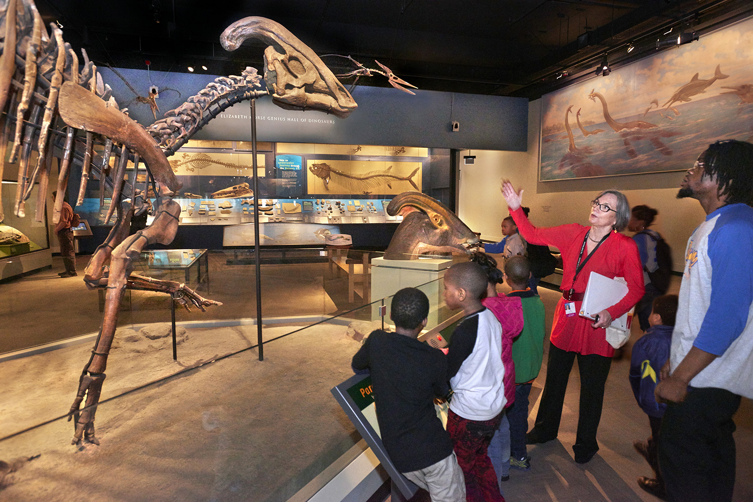 A Field Museum docent gestures to a Hadrosaurus skeleton while leading a tour group of students and teachers.