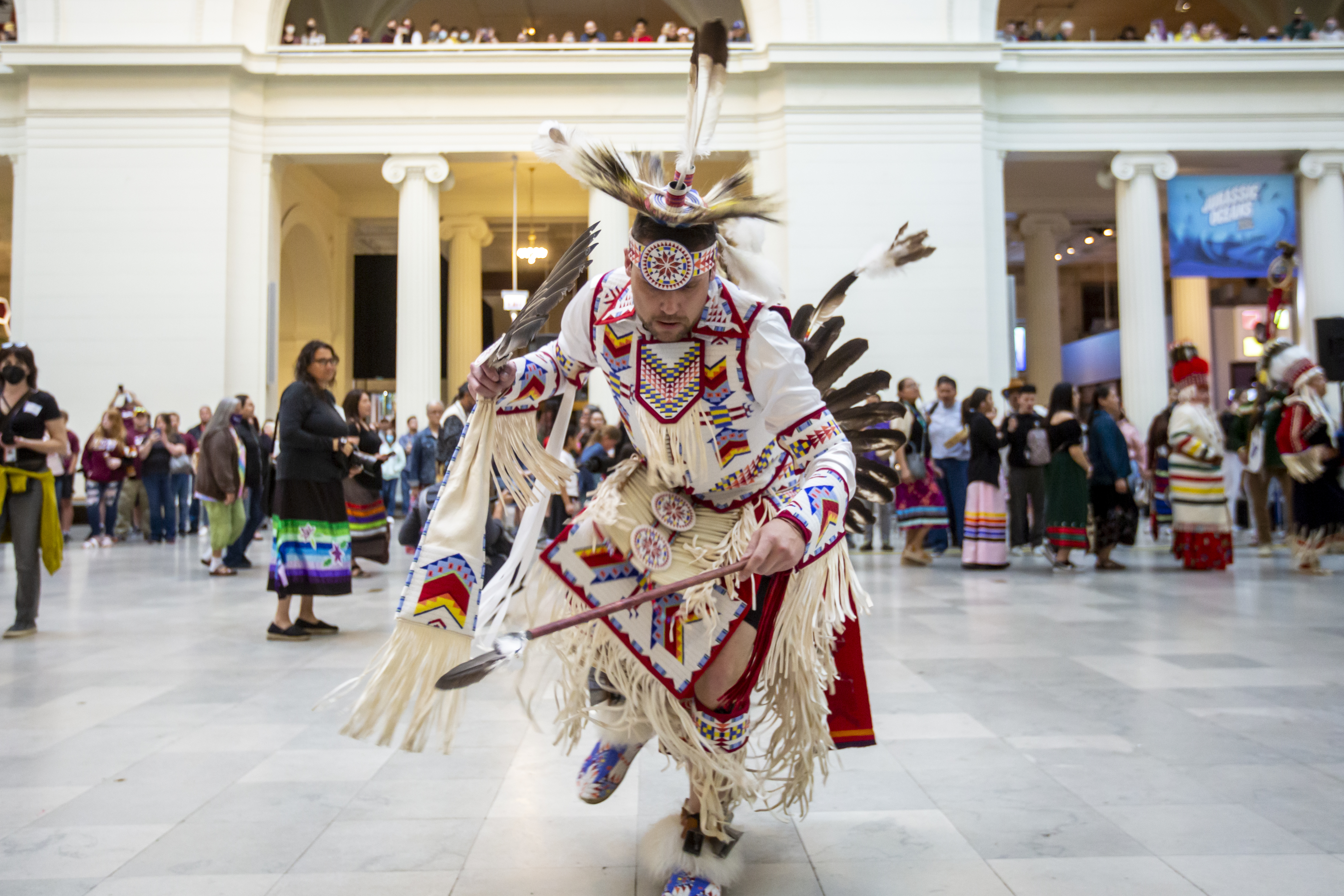 A Native American man wearing traditional regalia performs a stomp dance inside the Museum's main hall.