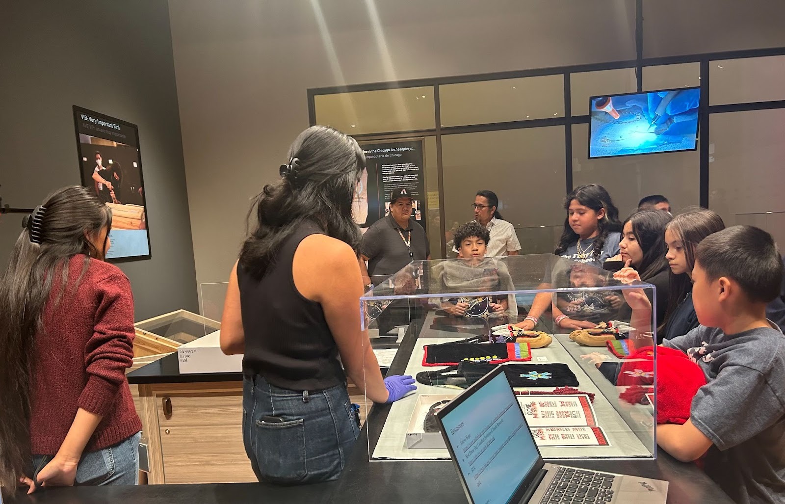 Two museum staff members present cultural items to a group of students standing on the other side of a counter.