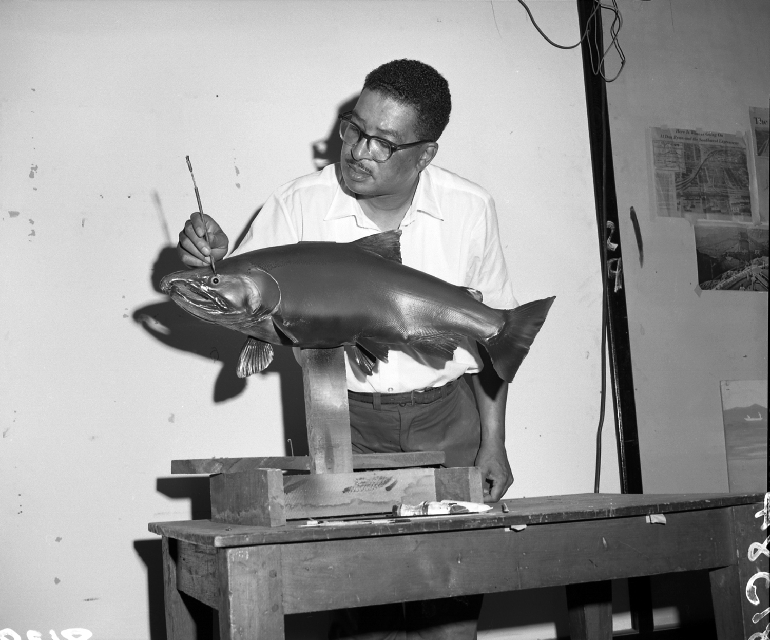 Carl Cotton using a long, thin tool to work on the eye detail of a salmon fish model. He wears a simple white collared shirt and horn-rimmed glasses and has a focused, patient expression.