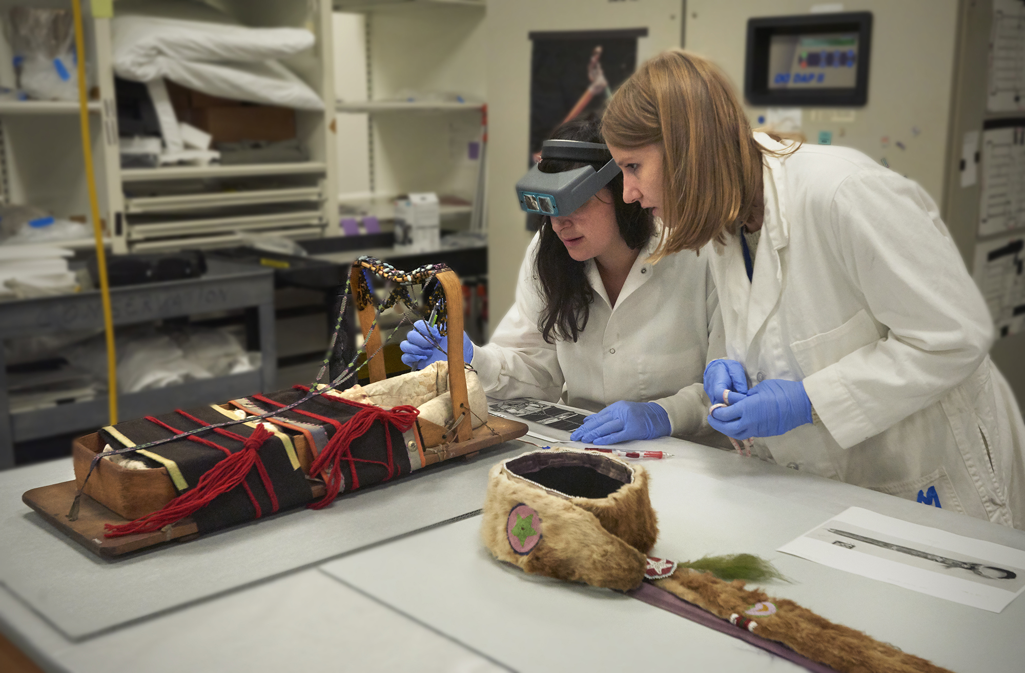 Two conservators testing a Native North American cradleboard in a lab.