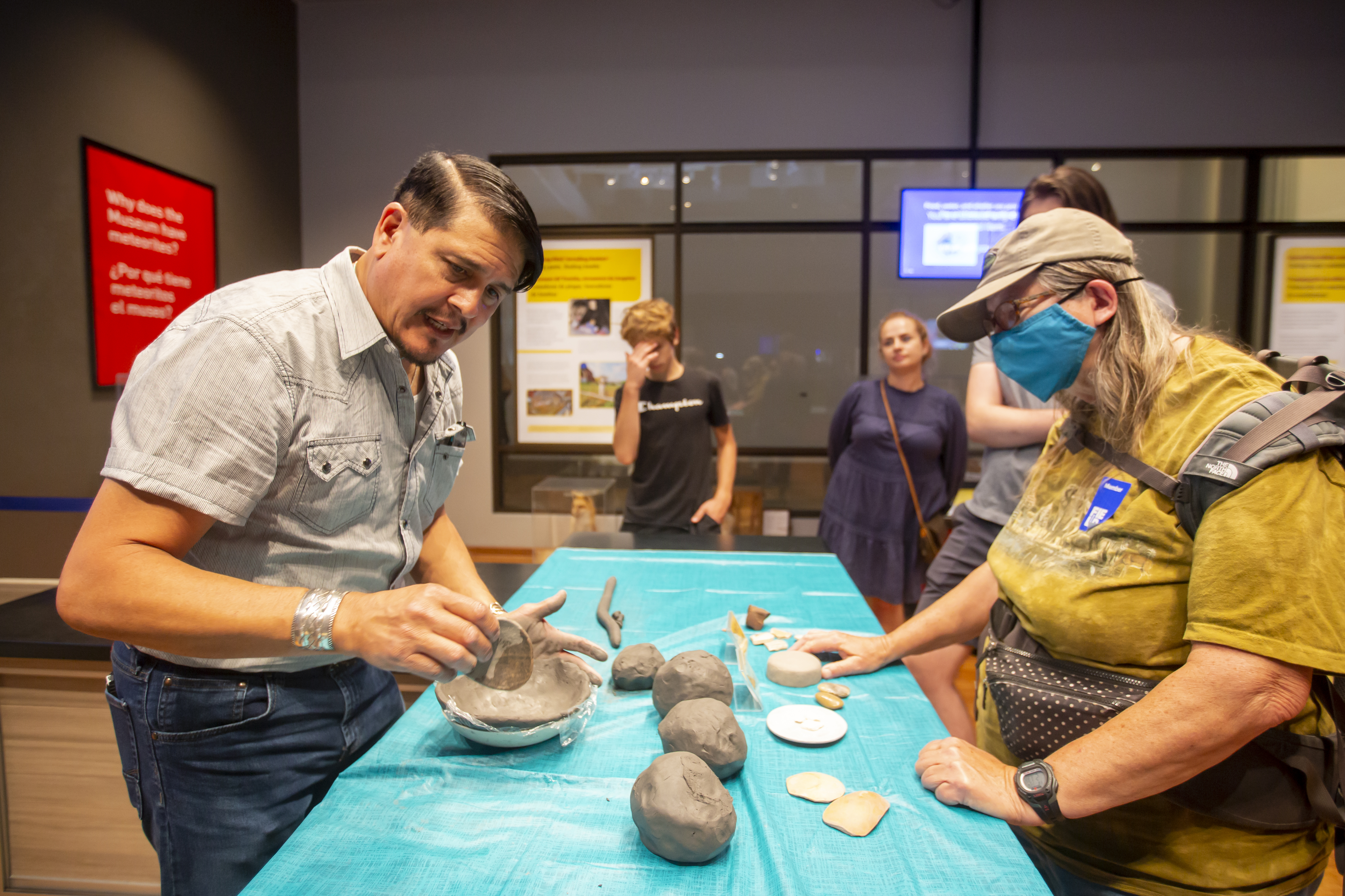 A pottery demonstration taking place in the Science Hub. A man and a woman stand on either side of a long table as the man gestures to a clay pot that he is molding. Lumps of clay and other tools are on the table in front of them.