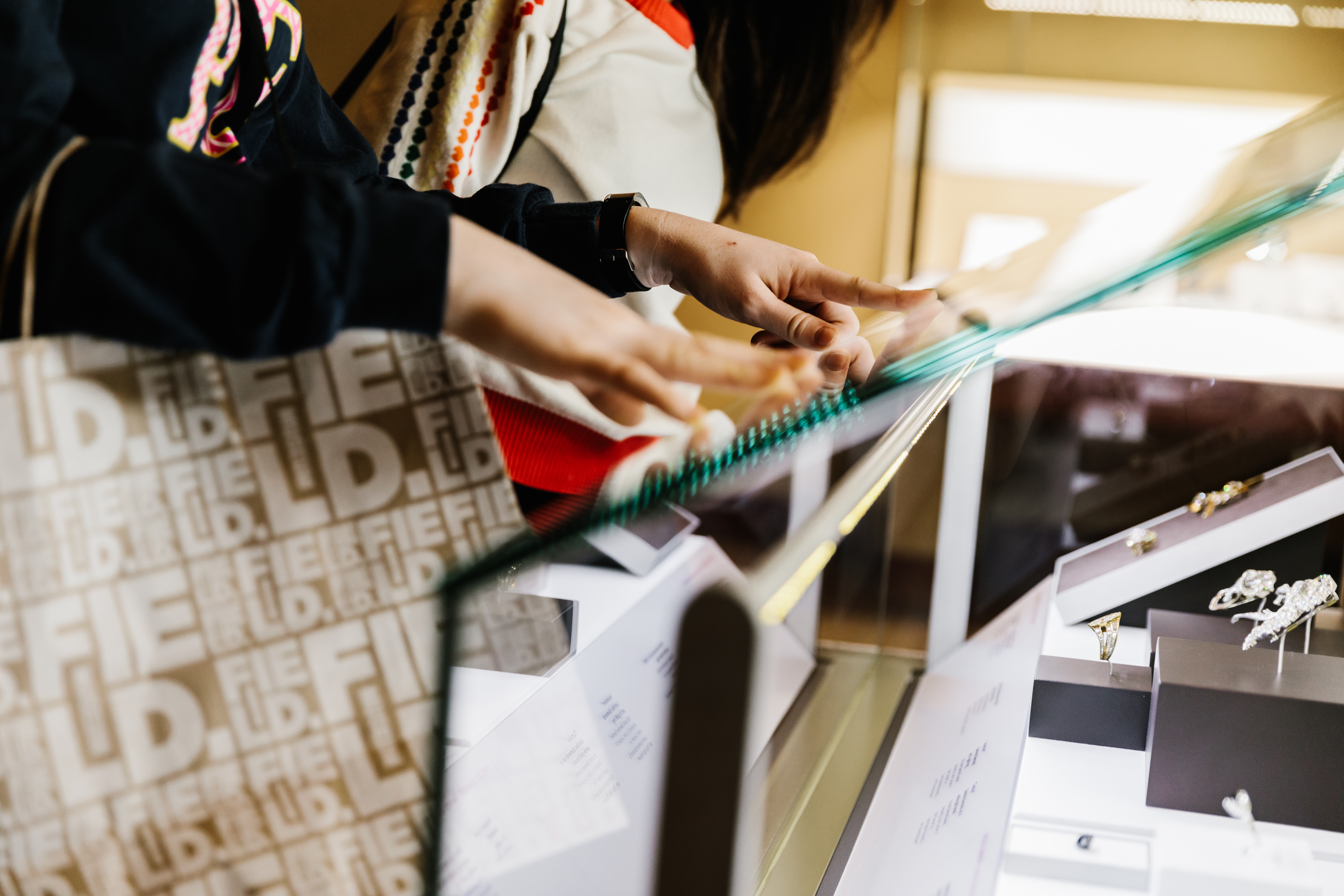 A visitor's hands on the glass case above sparkling ornaments in the Grainger Hall of Gems. The visitor has a Field Museum shopping bag hung over one arm. Another visitor stands nearby.