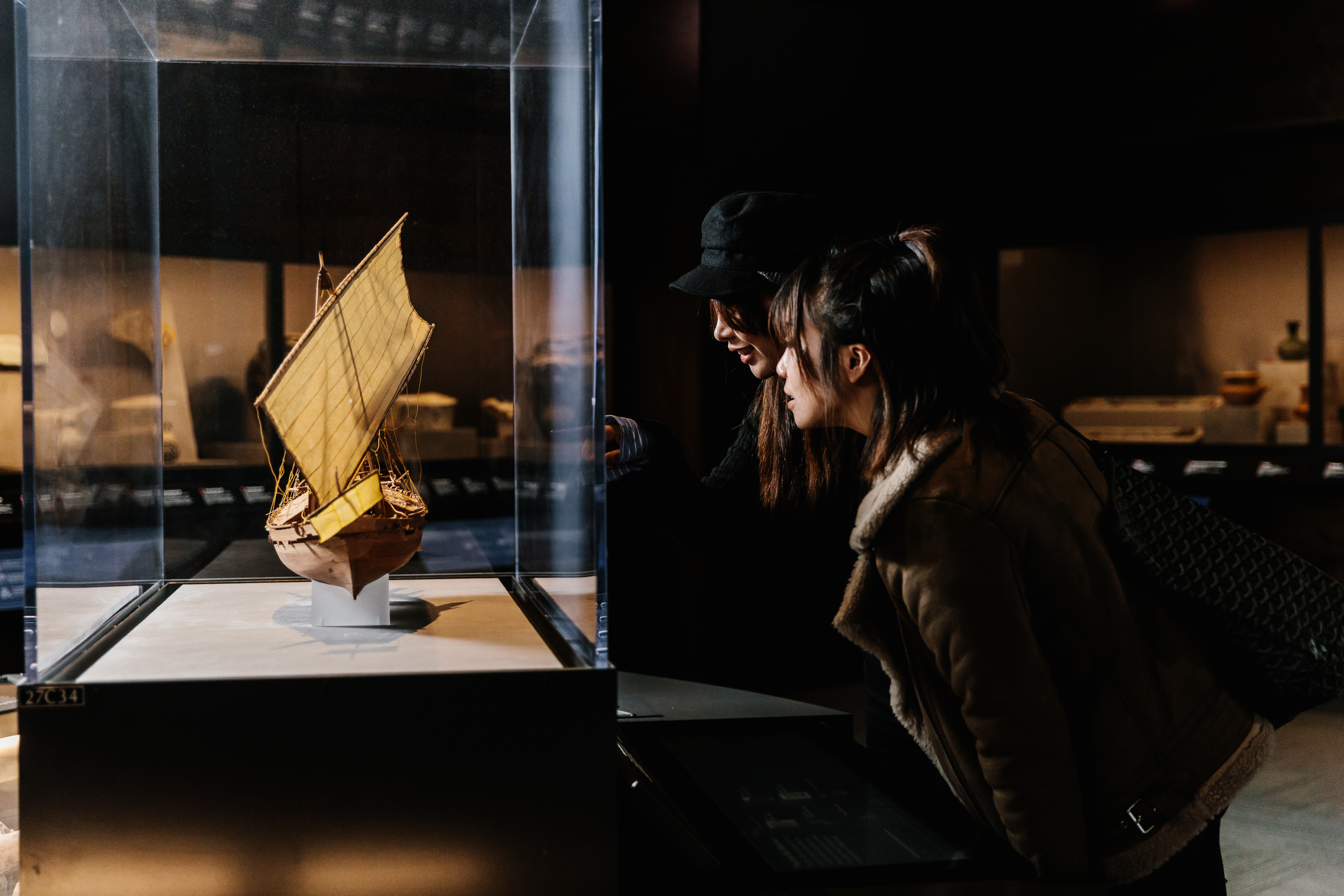 Two visitors observe a model of a ship in a glass case in a darkened gallery.