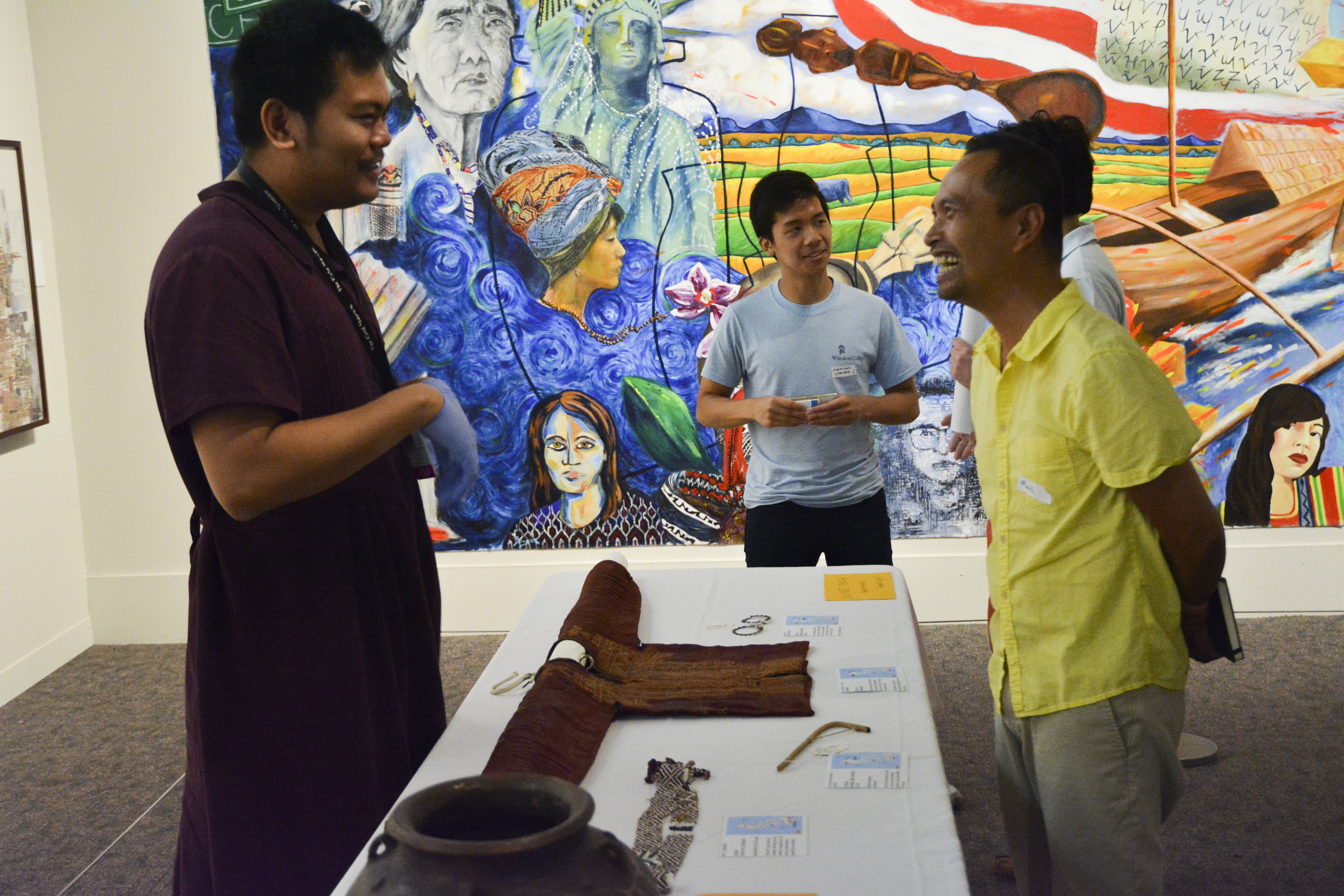 Two people talking over a woven shirt and pottery bowl. A large mural is in the background.