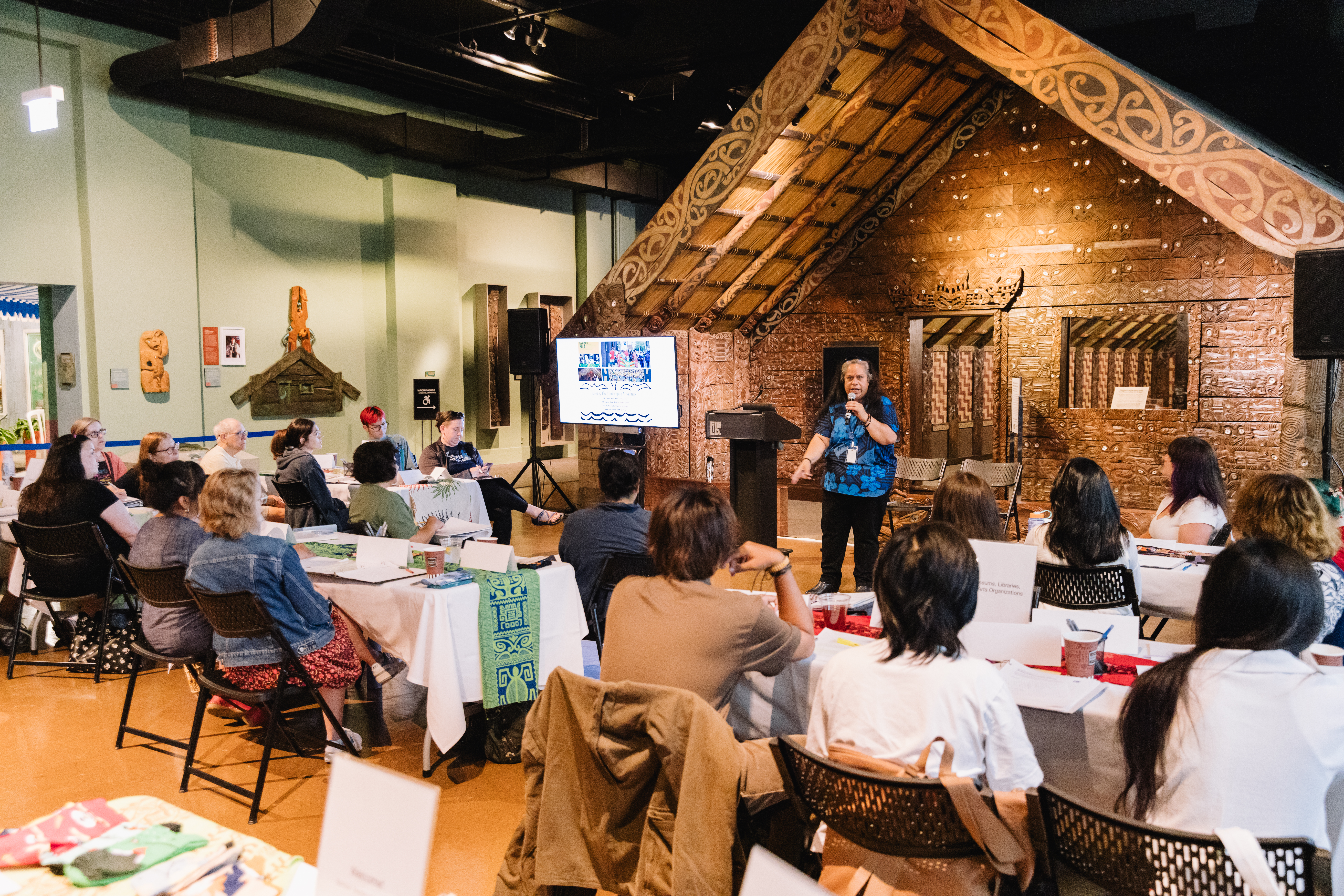 A group of adults seated at table, listening to a presentation in the Maori House gallery.