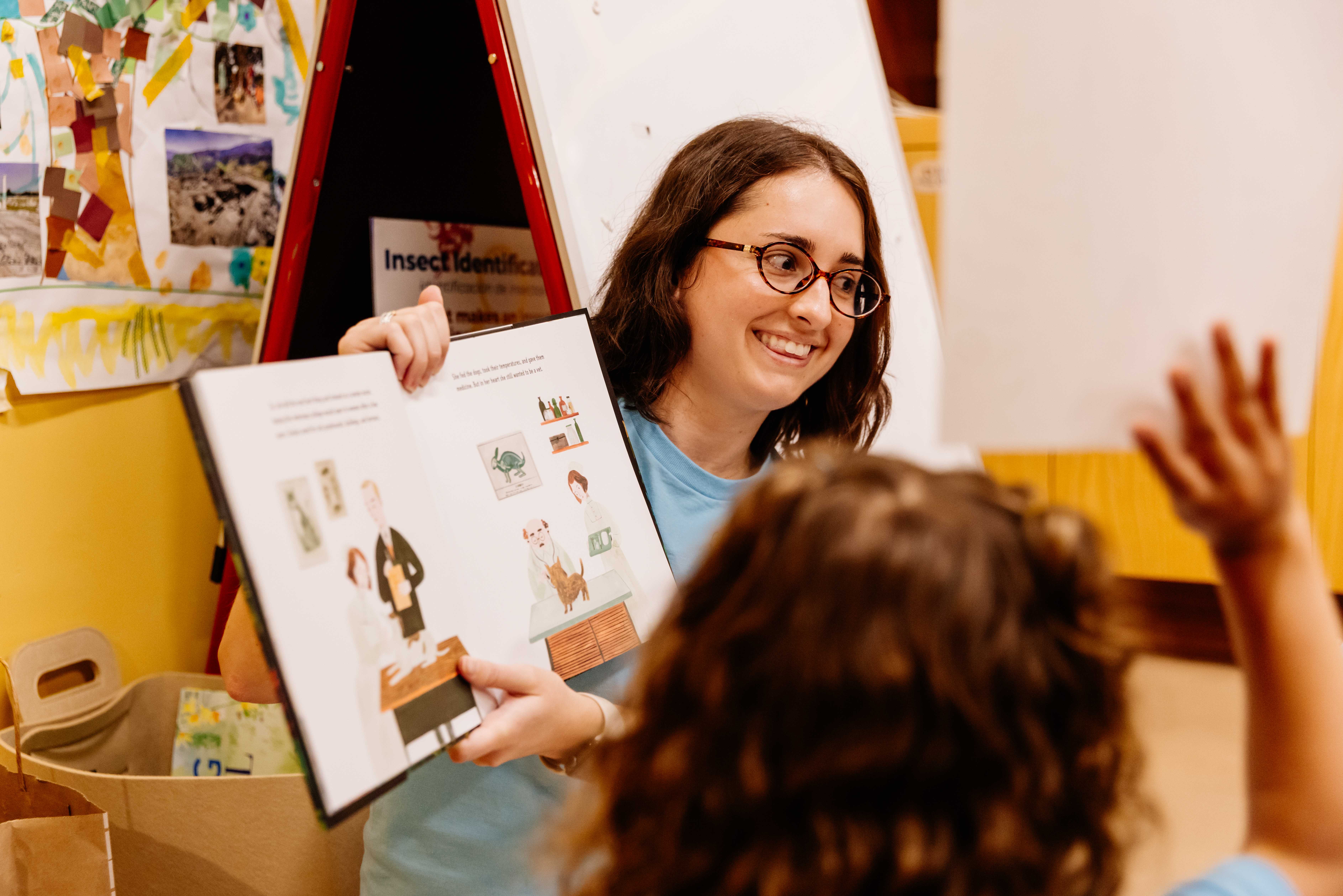 A woman holds an open book up to children during a story time event. The back of one child's head is visible in the foreground.