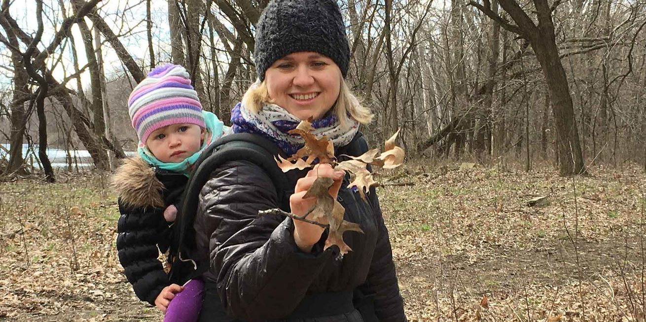 A mother and her young child explore the woods with a Field Museum field guide. The child, attached to her mother's back, looks on as her mother holds a bundle of dried leaves.